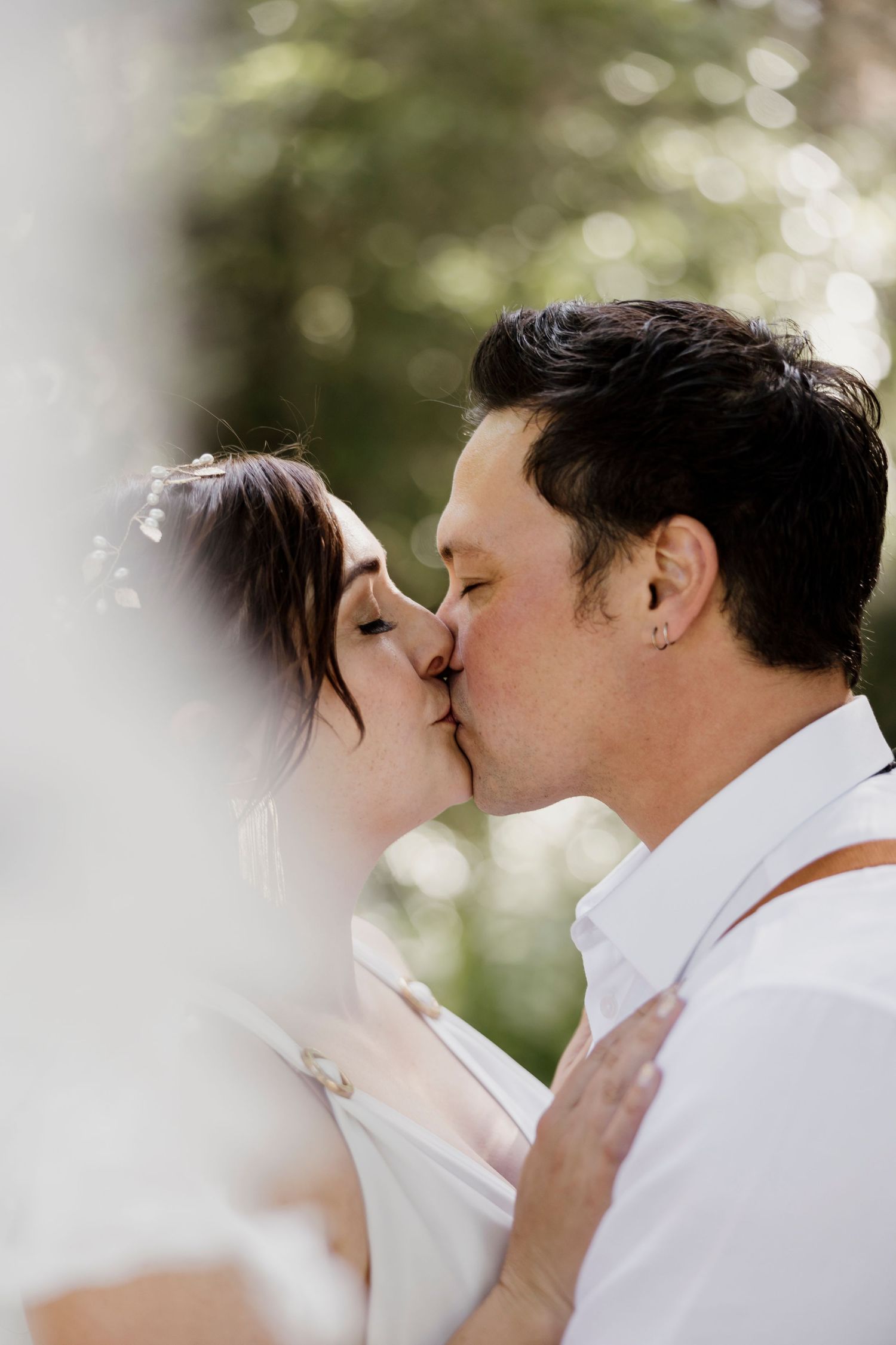 Bride and groom sharing their first kiss framed by soft bokeh greenery — Oh Joy! Photography Tauranga