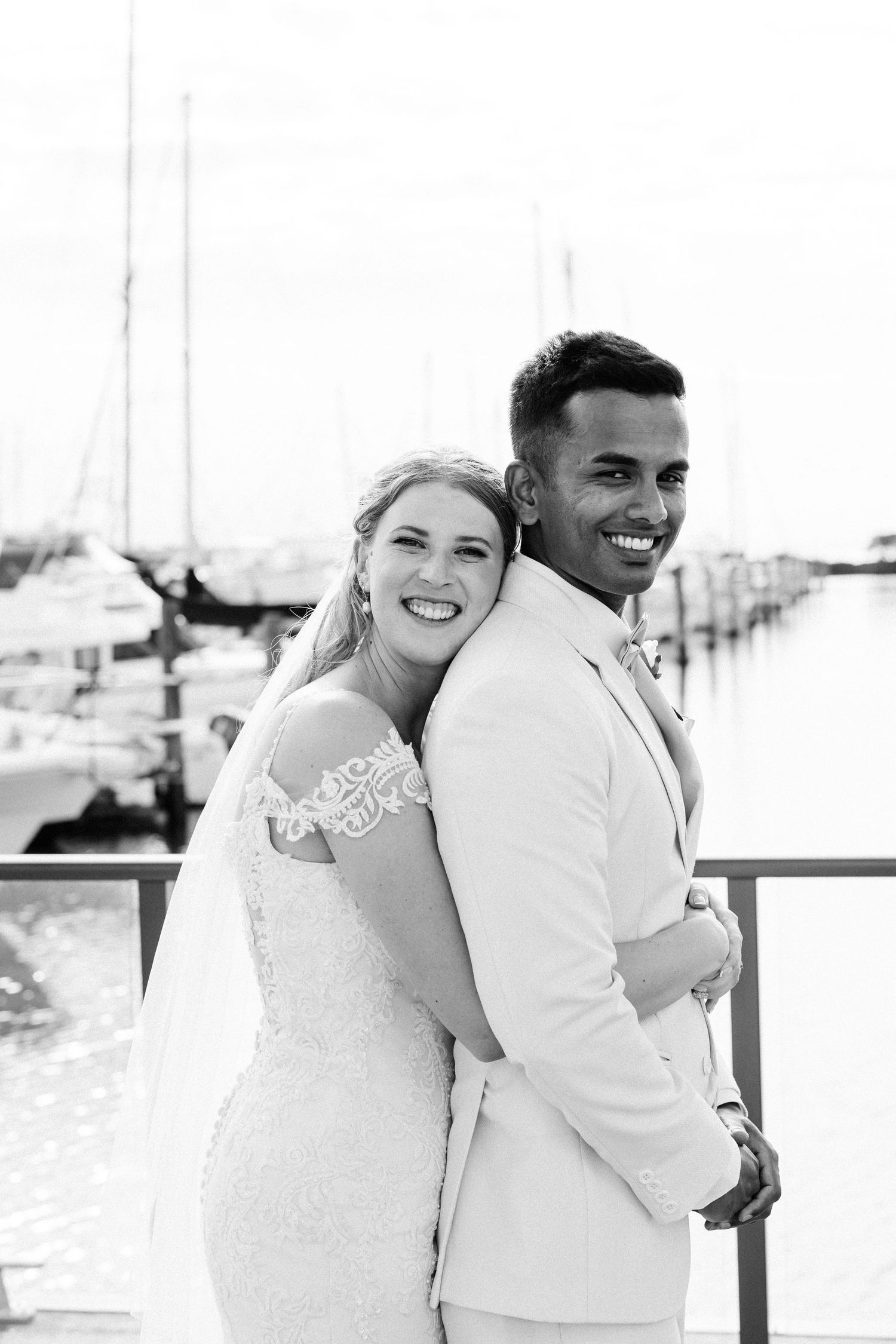 Black and white photo of laughing bride and groom back to back at a marina waterfront — Oh Joy! Photography Tauranga