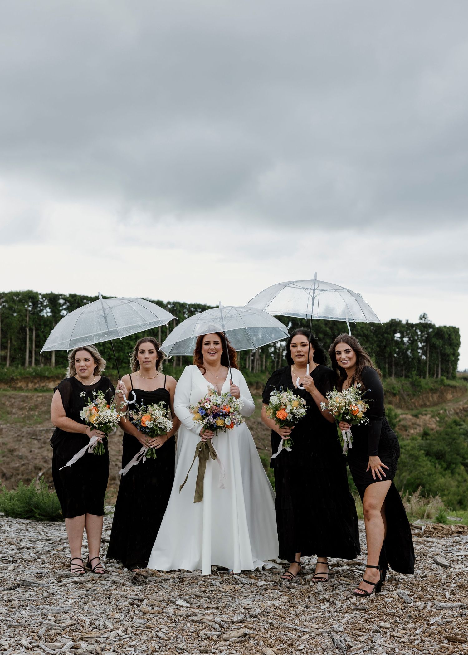 Bride and bridesmaids in black holding clear umbrellas and wildflower bouquets on a rainy NZ wedding day — Oh Joy! Photography