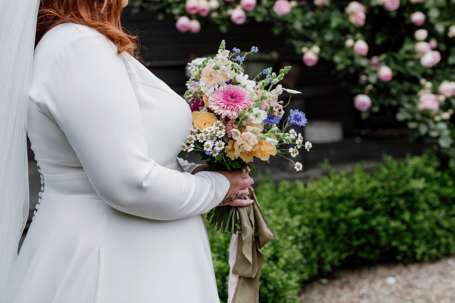 Bride in a long-sleeve white gown holding a colourful wildflower bouquet with roses and daisies — Oh Joy! Photography Tauranga