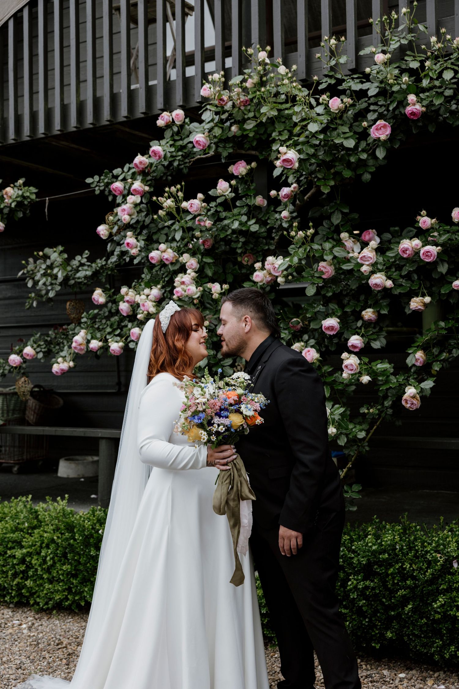 Bride and groom sharing an intimate moment before a climbing rose wall, wildflower bouquet in hand — Oh Joy! Photography Tauranga