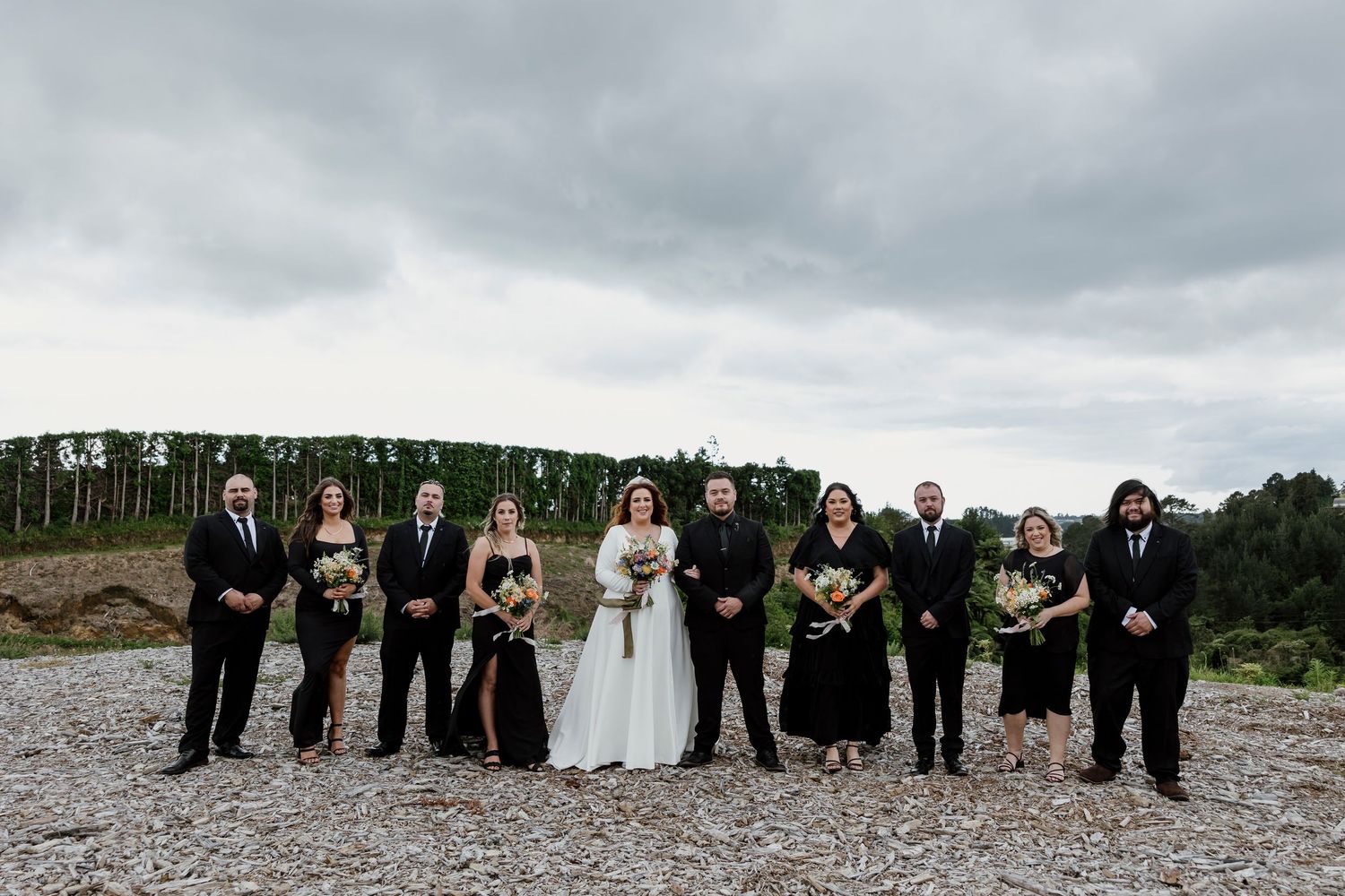 Bride with wedding party in black, holding wildflower bouquets against a dramatic NZ landscape — Oh Joy! Photography Tauranga