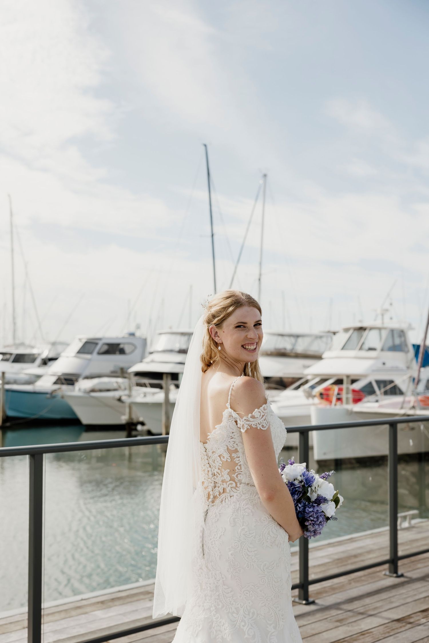 Smiling bride in a lace off-shoulder gown holding a purple bouquet at a sunny marina — Oh Joy! Photography Tauranga