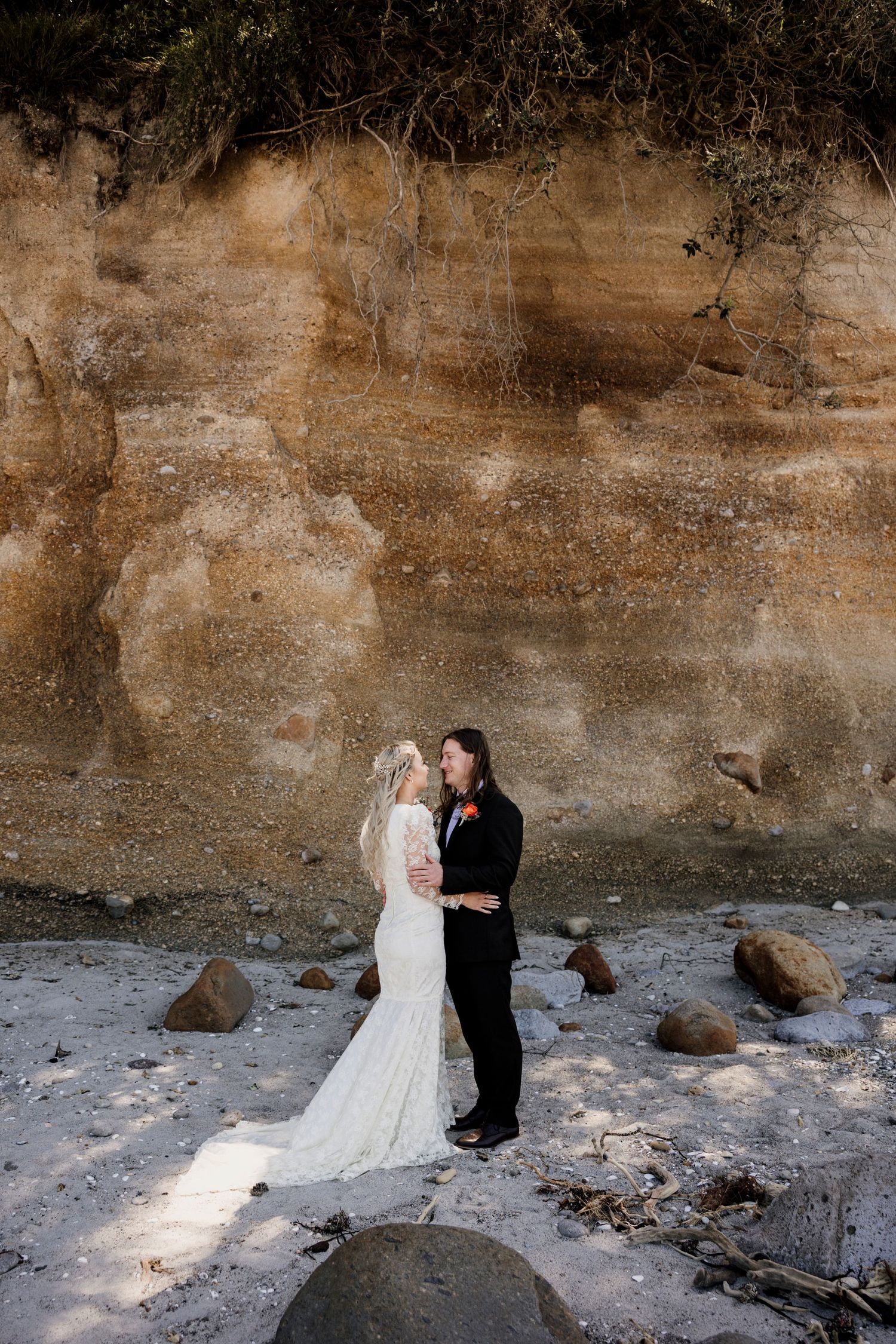 Bride and groom sharing a joyful moment on a rocky NZ beach with dramatic sandstone cliffs — Oh Joy! Photography Tauranga