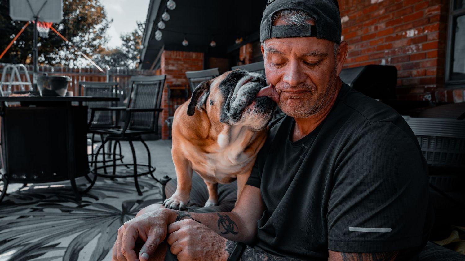 Man with tattoos cuddles an English Bulldog on an outdoor patio.