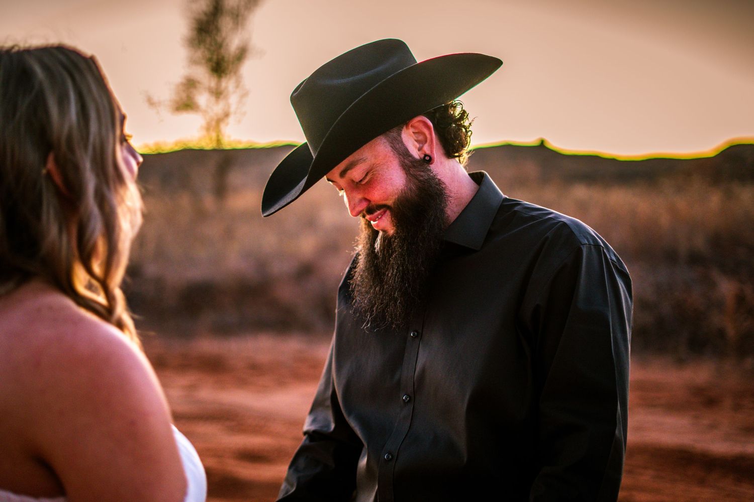 Bearded groom in black cowboy hat smiling warmly during an outdoor sunset ceremony.