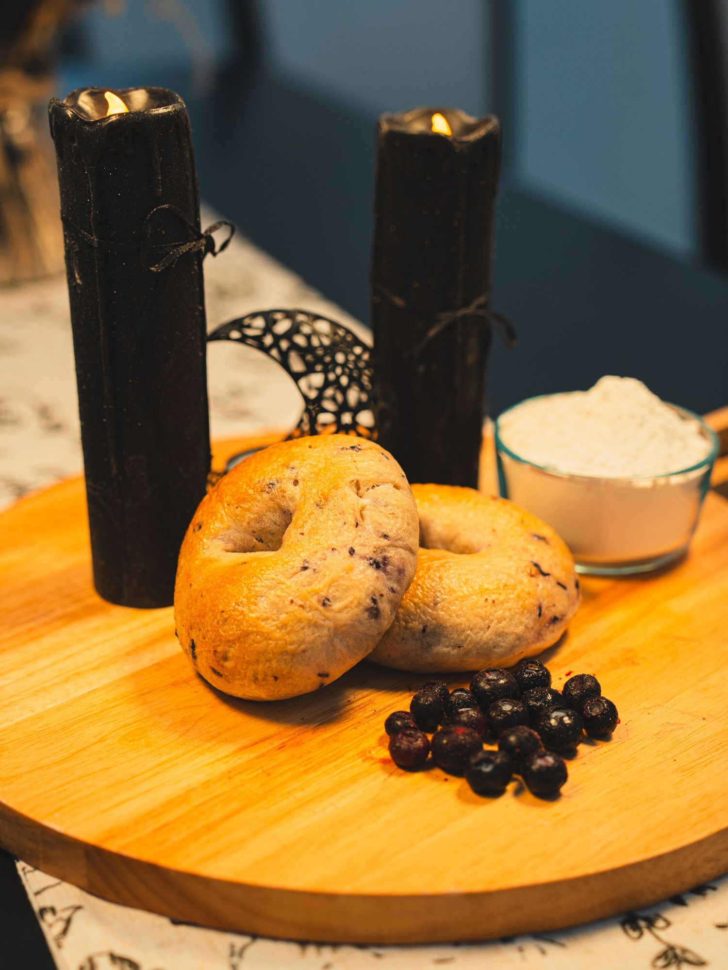 Blueberry bagels on a wooden board with fresh blueberries, cream cheese, and black candles in the background.