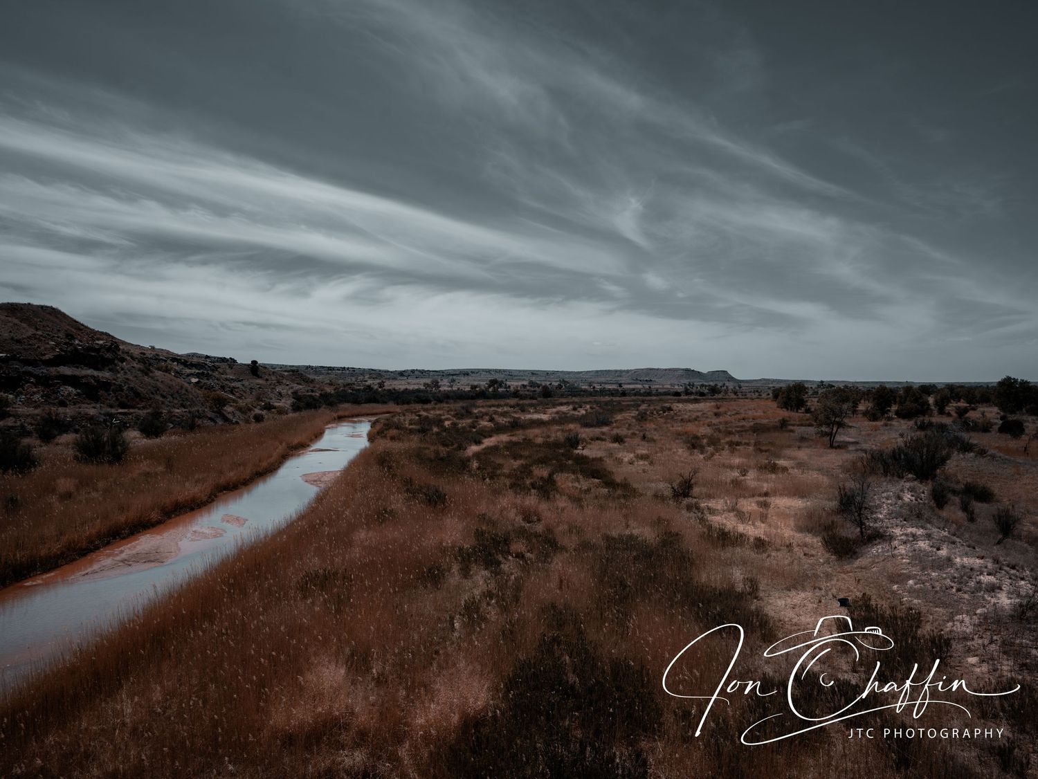 A winding river cuts through a dry golden grassland with red rock formations under a dramatic cloudy sky.