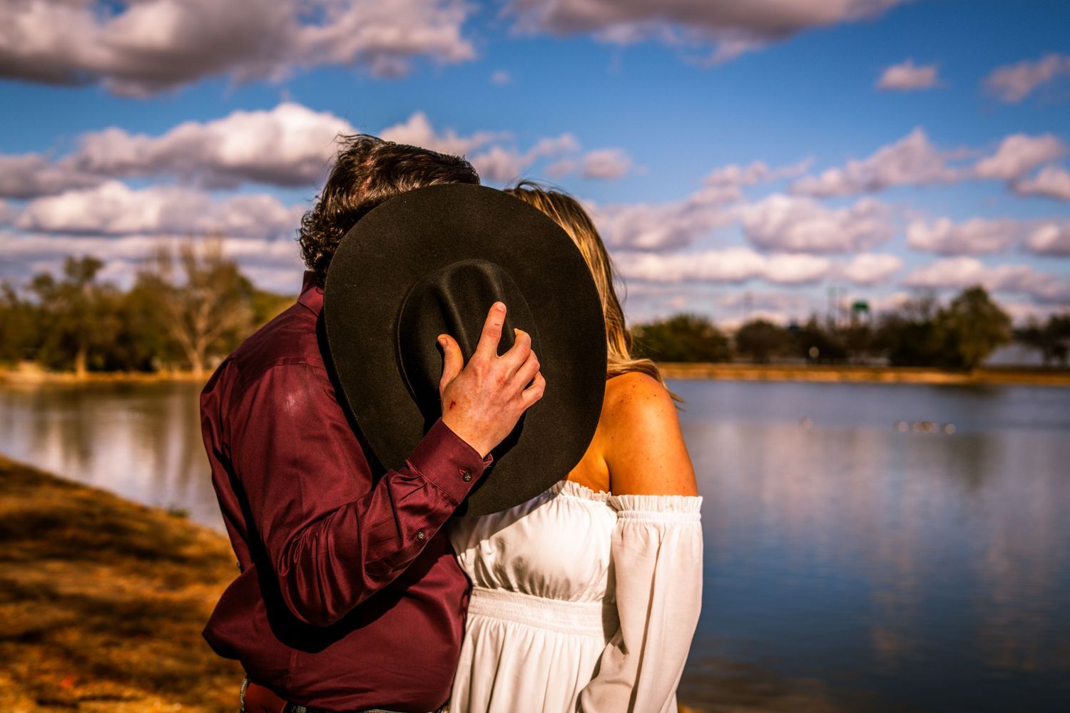 Romantic couple kissing behind a black cowboy hat near a sunlit lake with dramatic cloudy blue sky.
