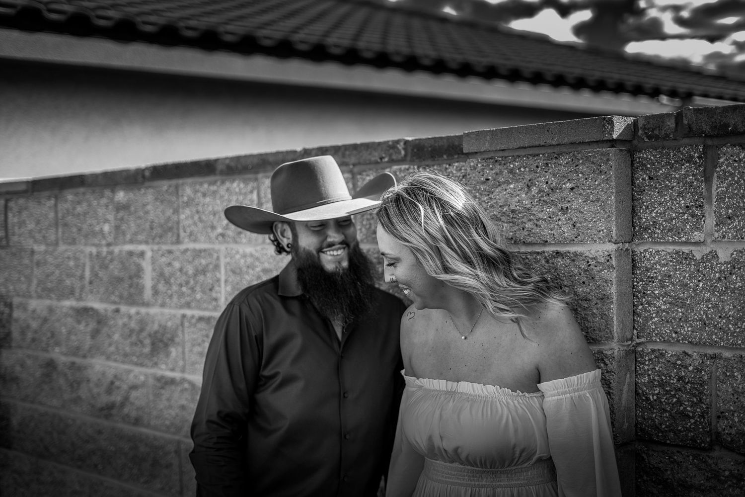 Black and white romantic portrait of a bearded man in a cowboy hat and woman leaning against a stone wall.