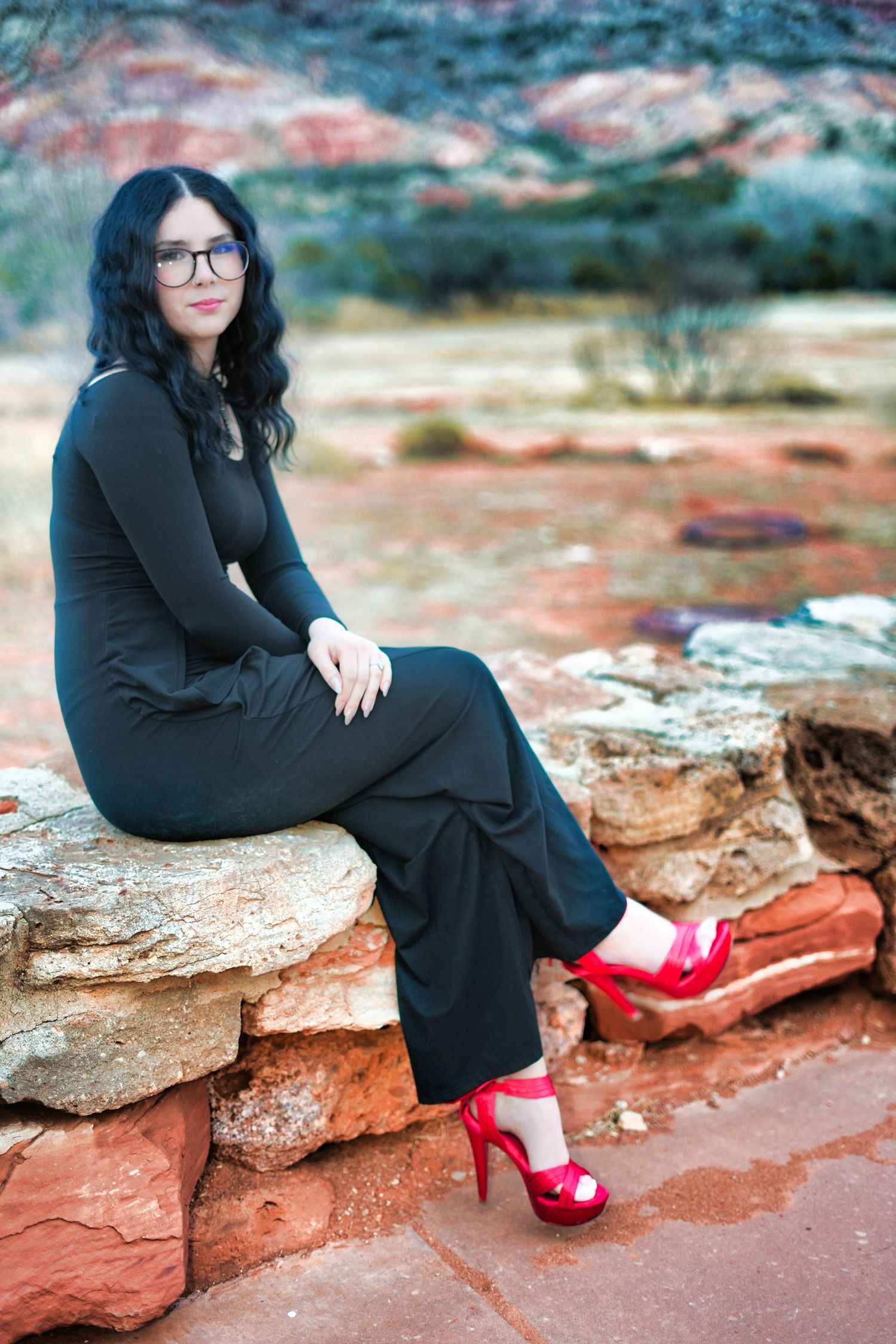 Woman in black outfit and red heels sitting on stone wall in red rock desert.
