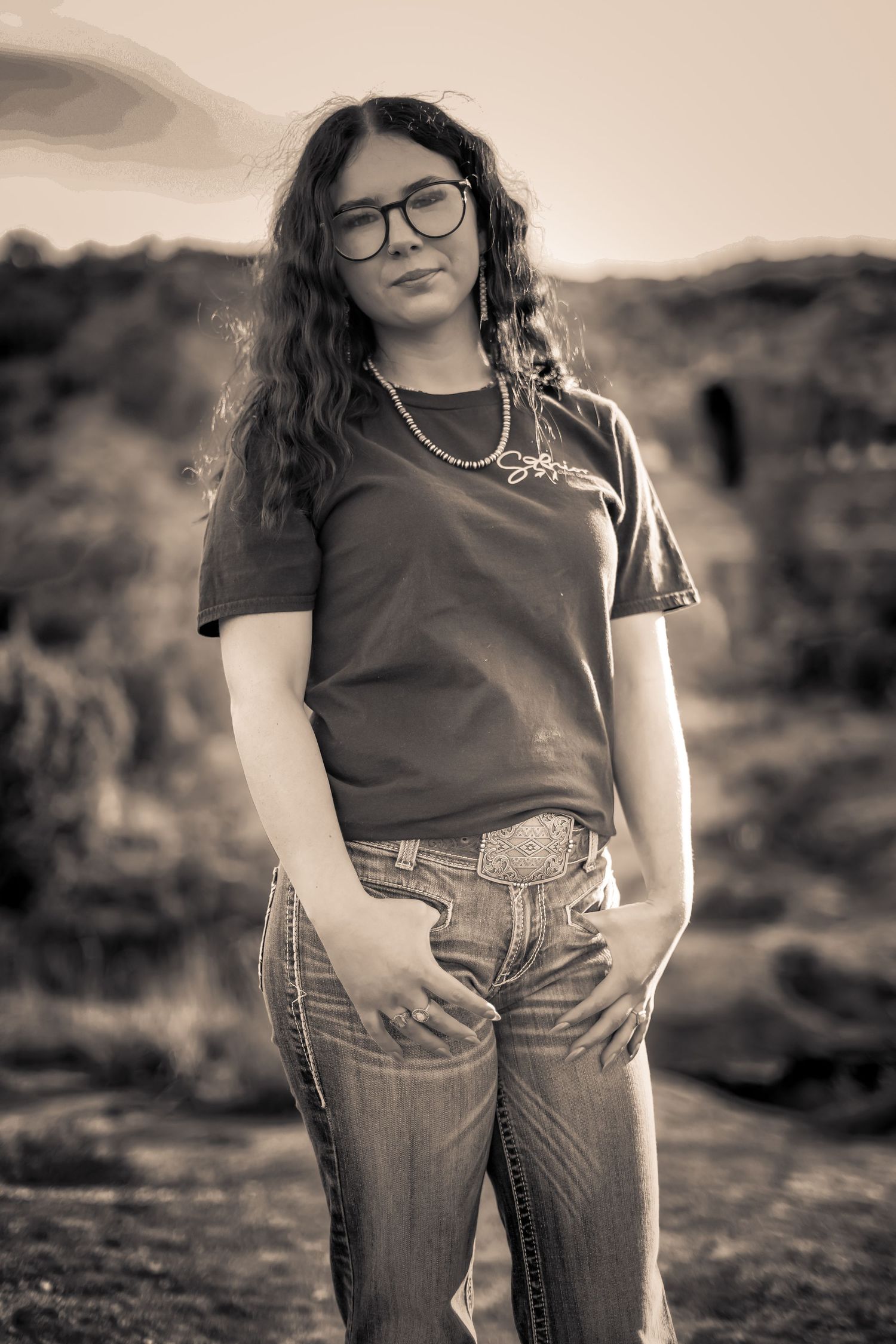 Young woman in glasses, jeans, and graphic tee poses outdoors in sepia-toned desert portrait.