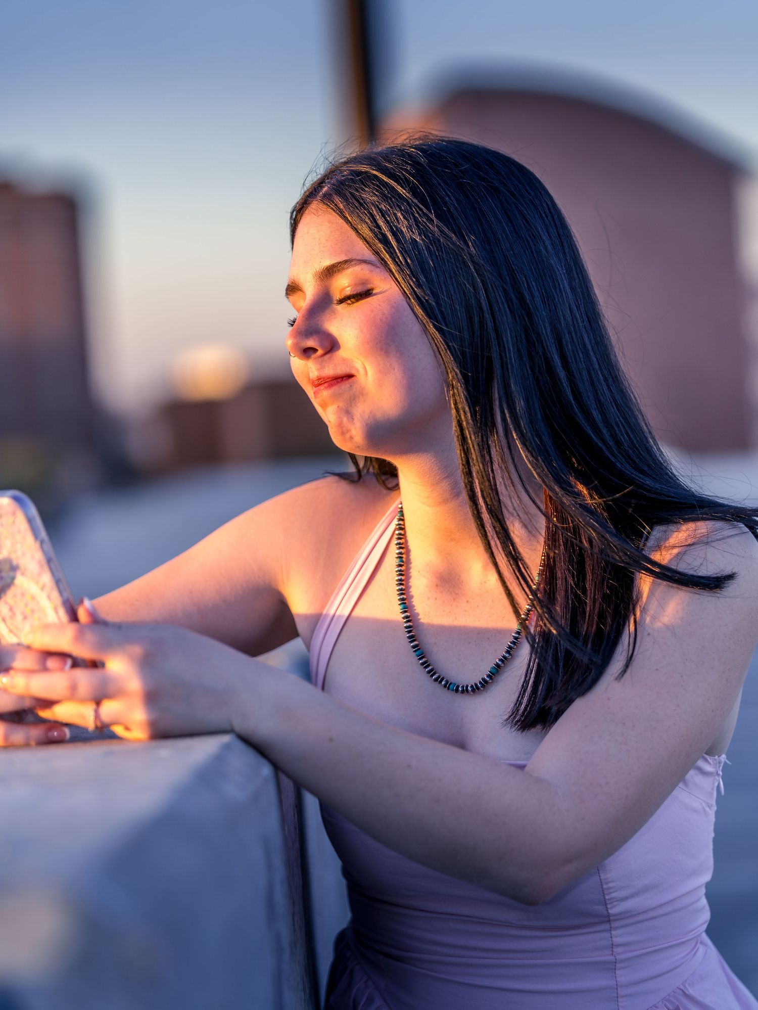 Dark-haired woman in white dress leans on wall during golden sunset.