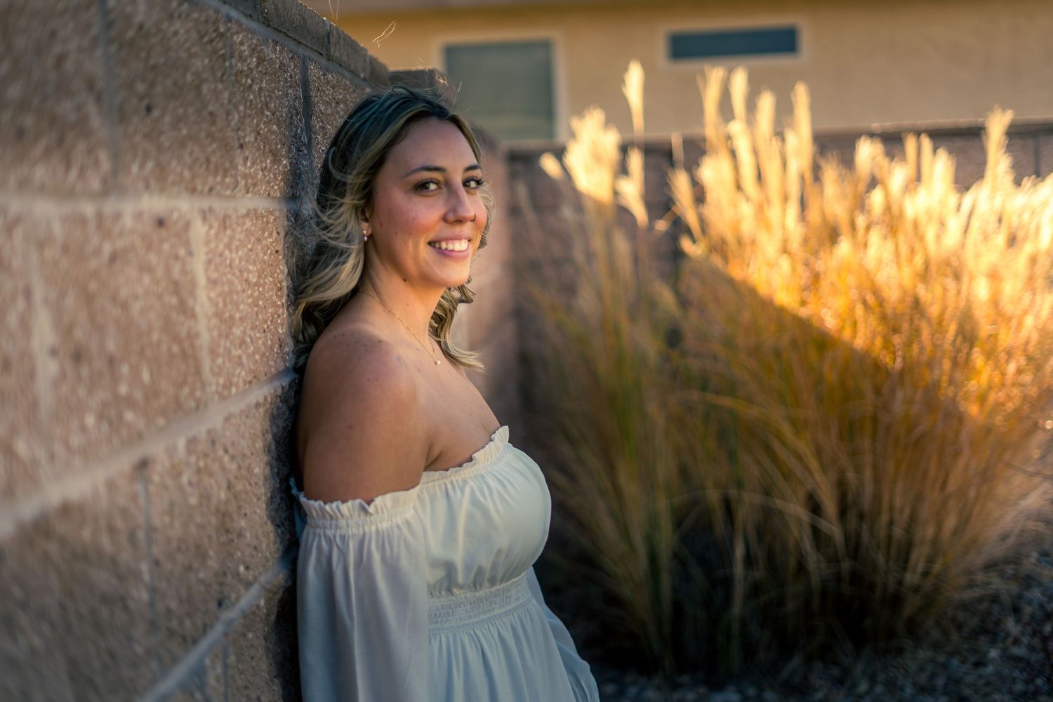 Young woman in white off-shoulder dress smiling against a stone wall at golden hour.