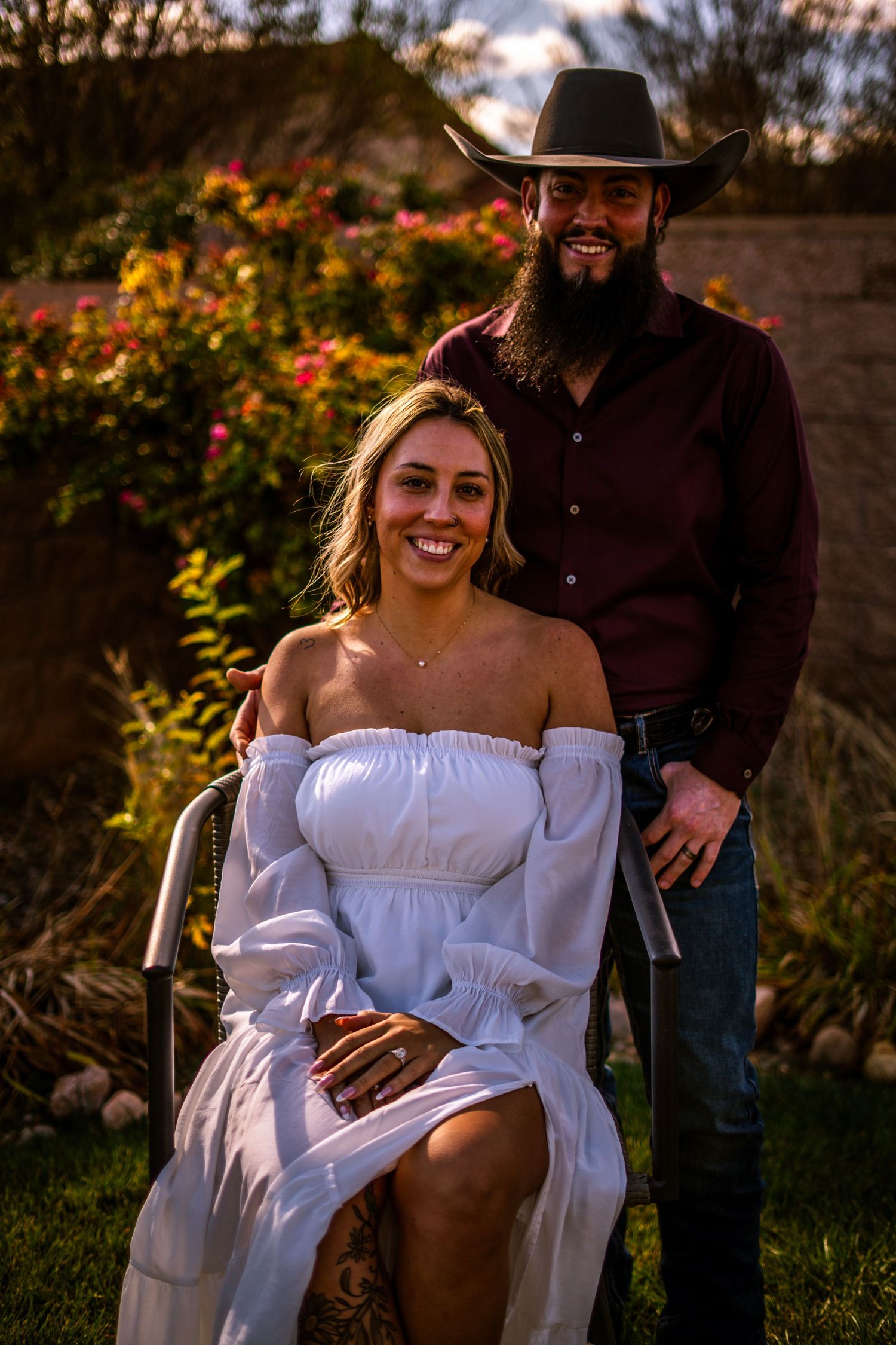 Smiling couple in garden, woman in white dress seated as bearded man in cowboy hat and maroon shirt stands behind.