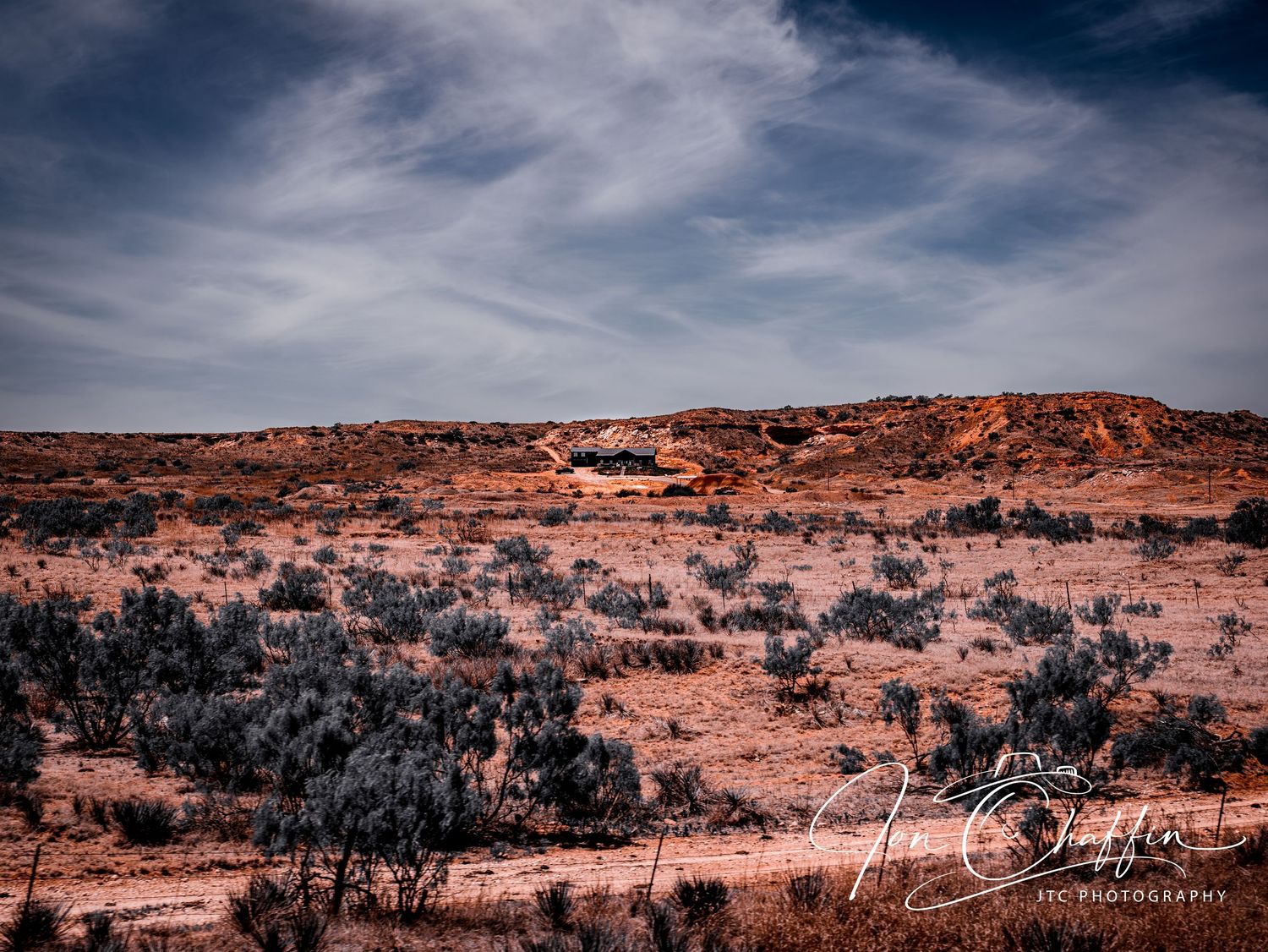 Arid desert landscape with sparse shrubs and a lone structure under cloudy sky.
