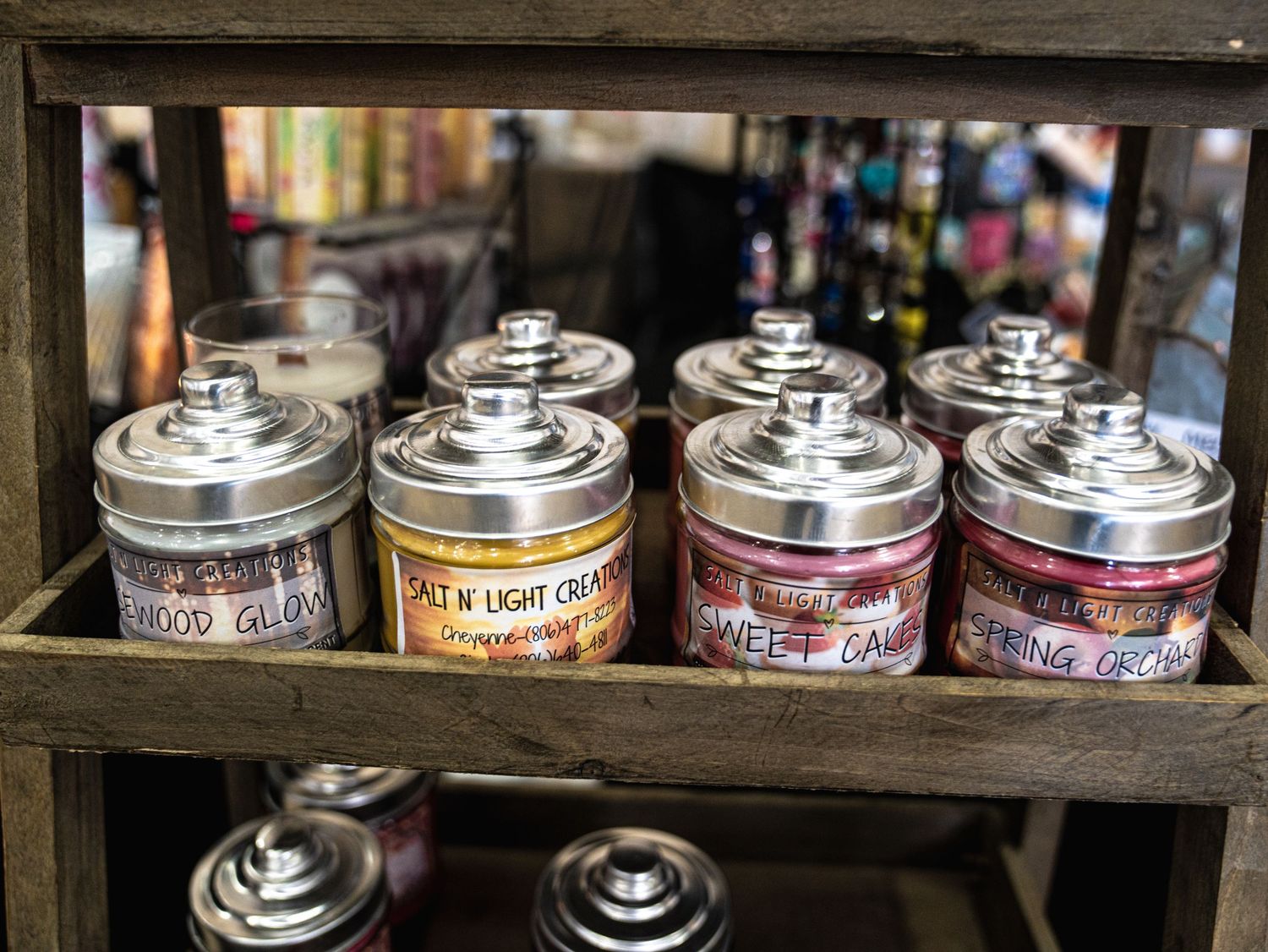 Assorted scented candles in silver-lidded glass jars displayed on a rustic wooden shelf in a gift shop.
