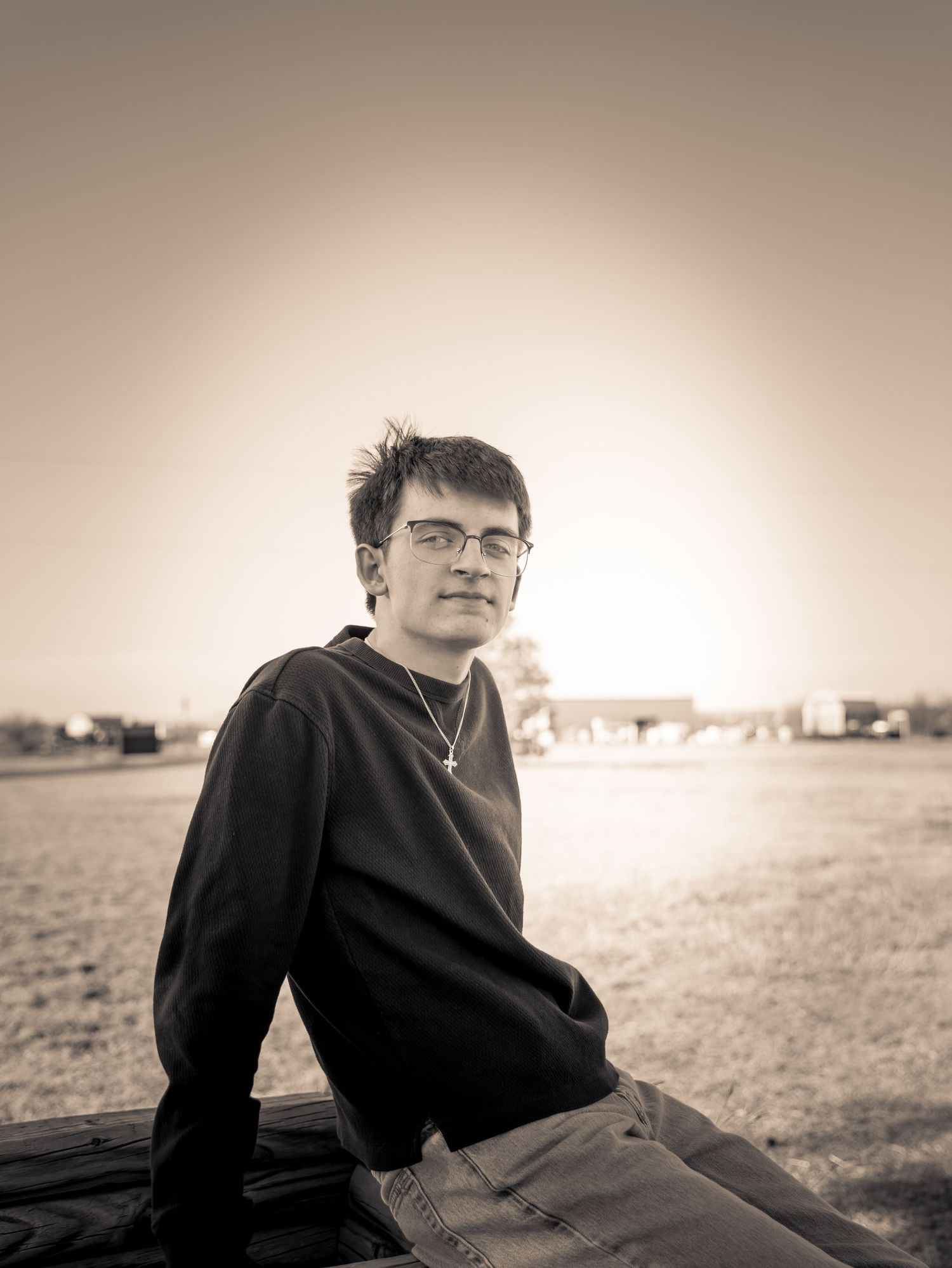 Black and white portrait of a young person with glasses sitting outdoors at sunset wearing a cross necklace.