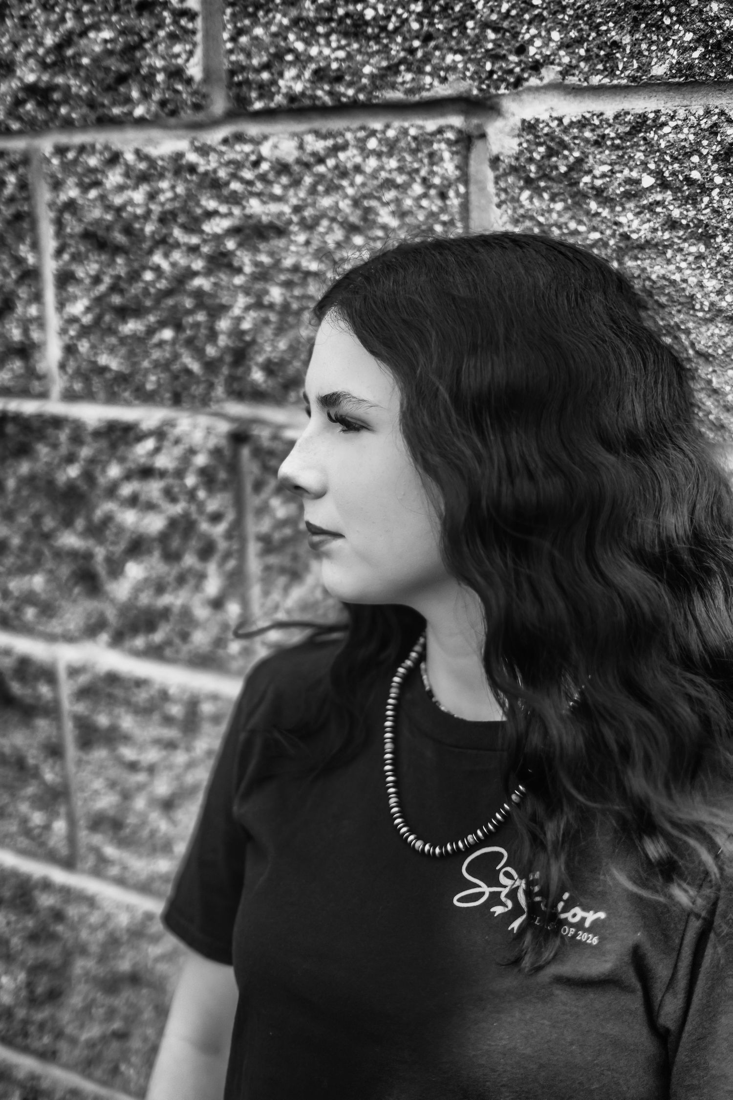 Black and white close-up portrait of a young woman with wavy dark hair gazing sideways against a stone wall.