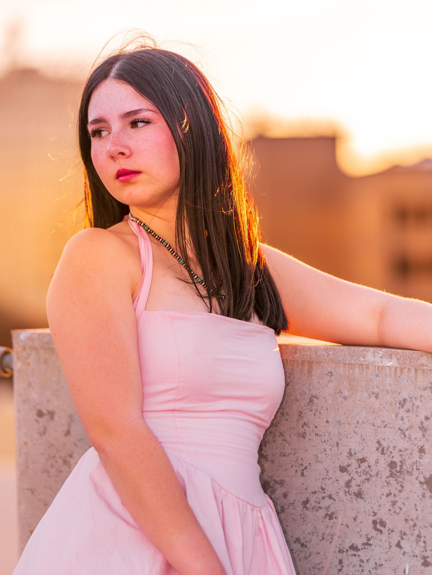 Young woman in pink dress leaning against concrete wall during golden sunset outdoors.