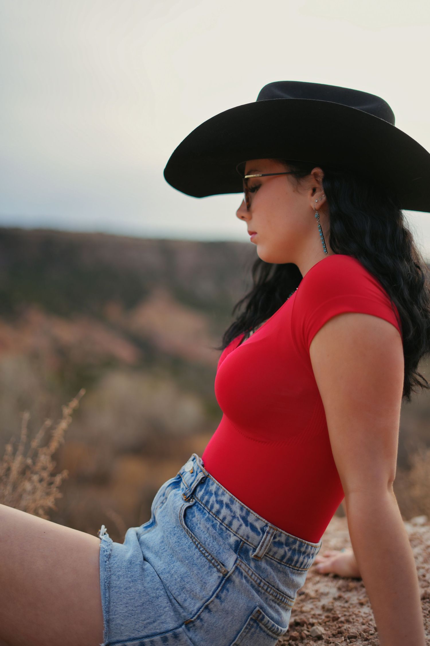 Young woman in black cowboy hat and red top sits on rocky terrain with canyon views.