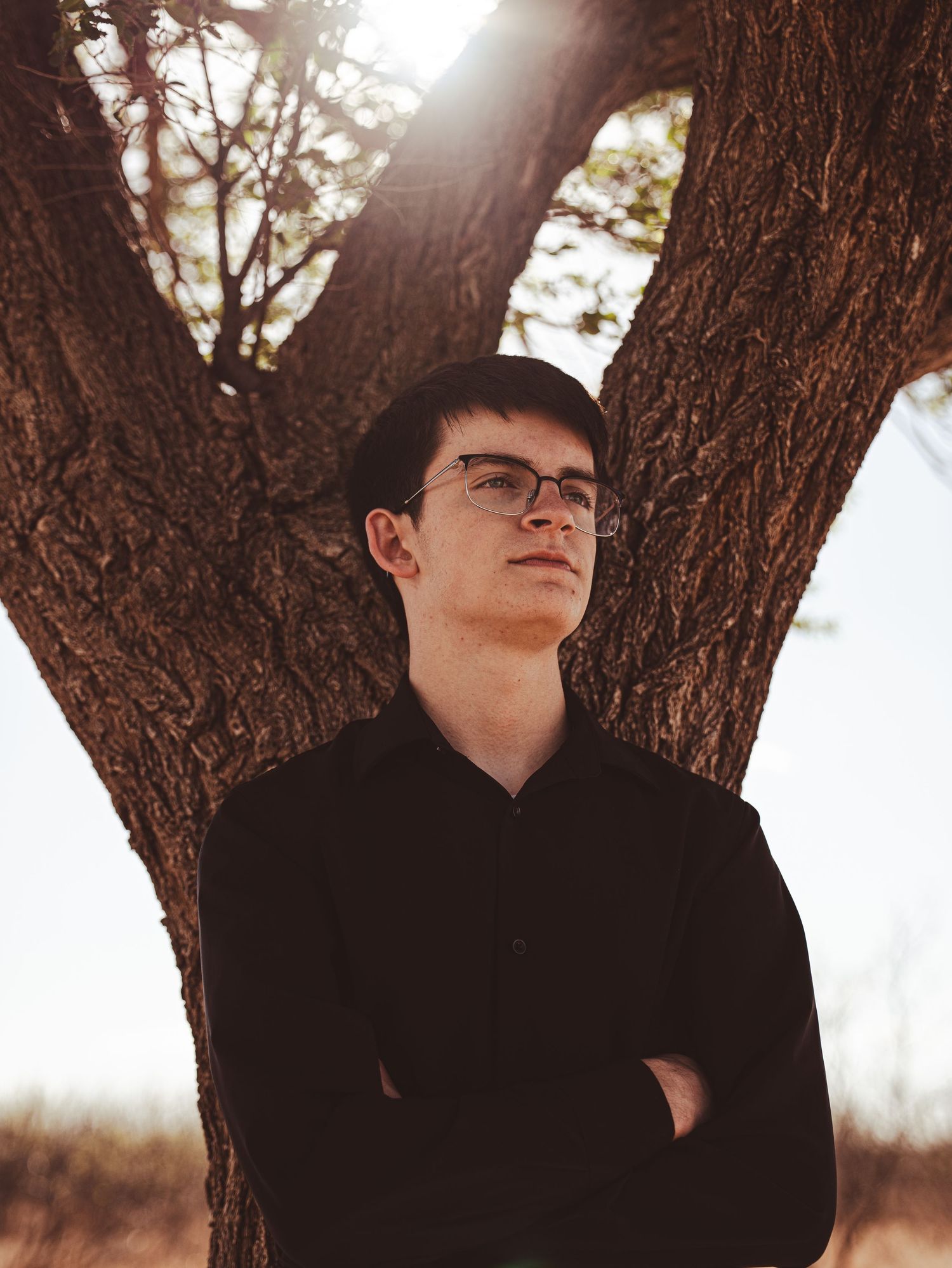Young man in black shirt with crossed arms standing against a large tree in golden light.