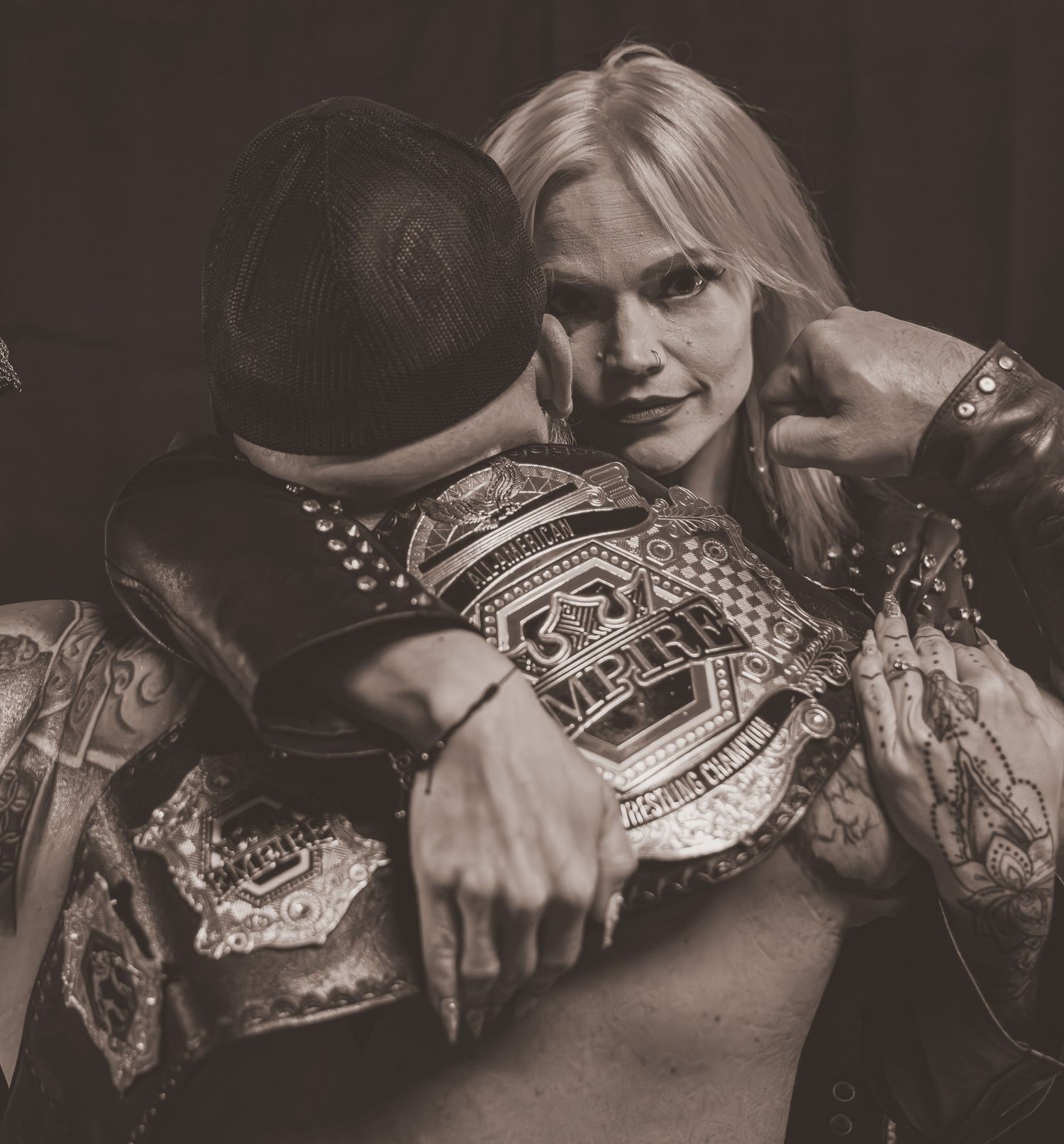 Sepia-toned photo of blonde female wrestler hugging tattooed man while clutching championship belt.