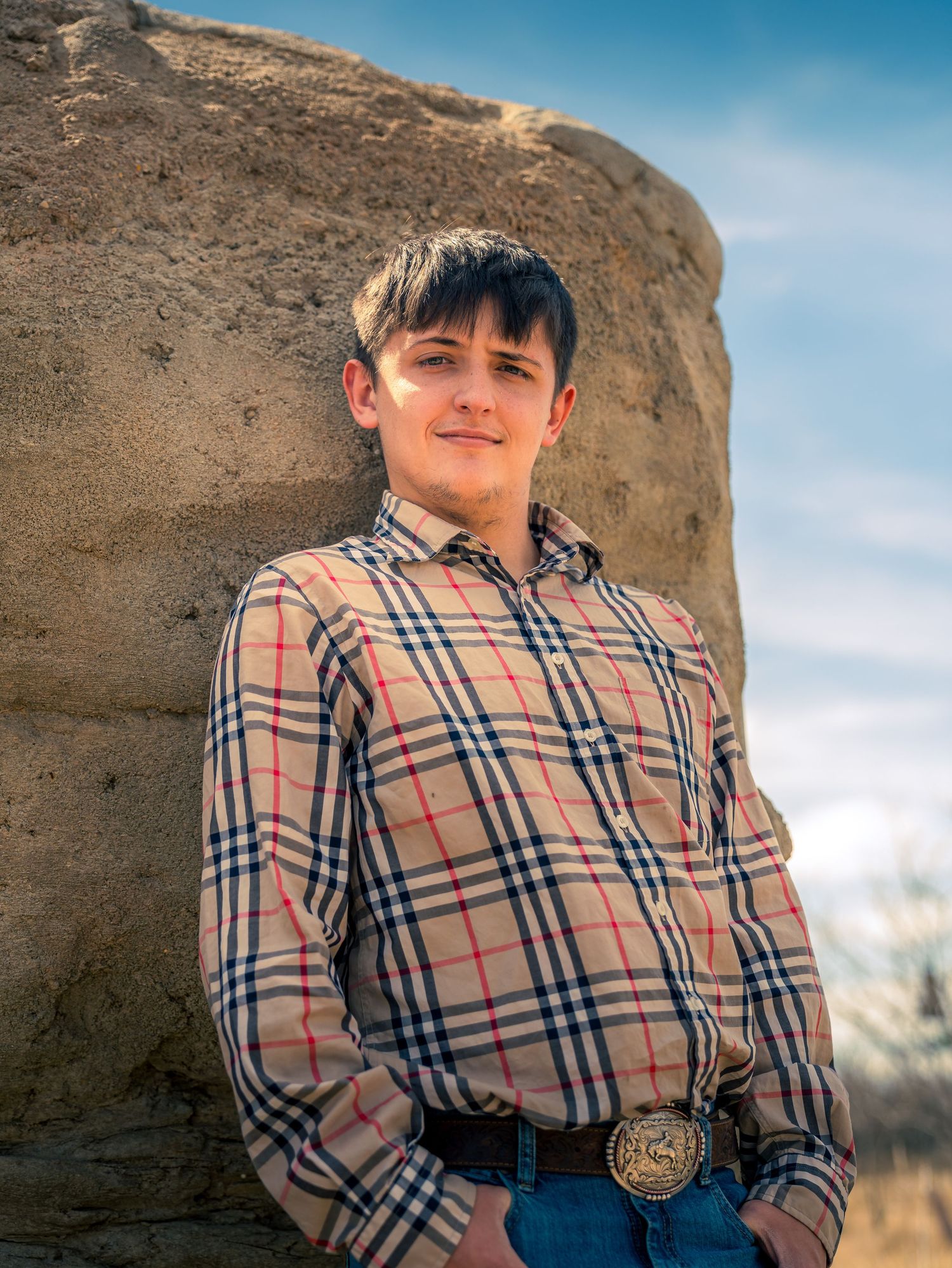 Young man in plaid shirt and belt buckle leaning against a rock formation outdoors.