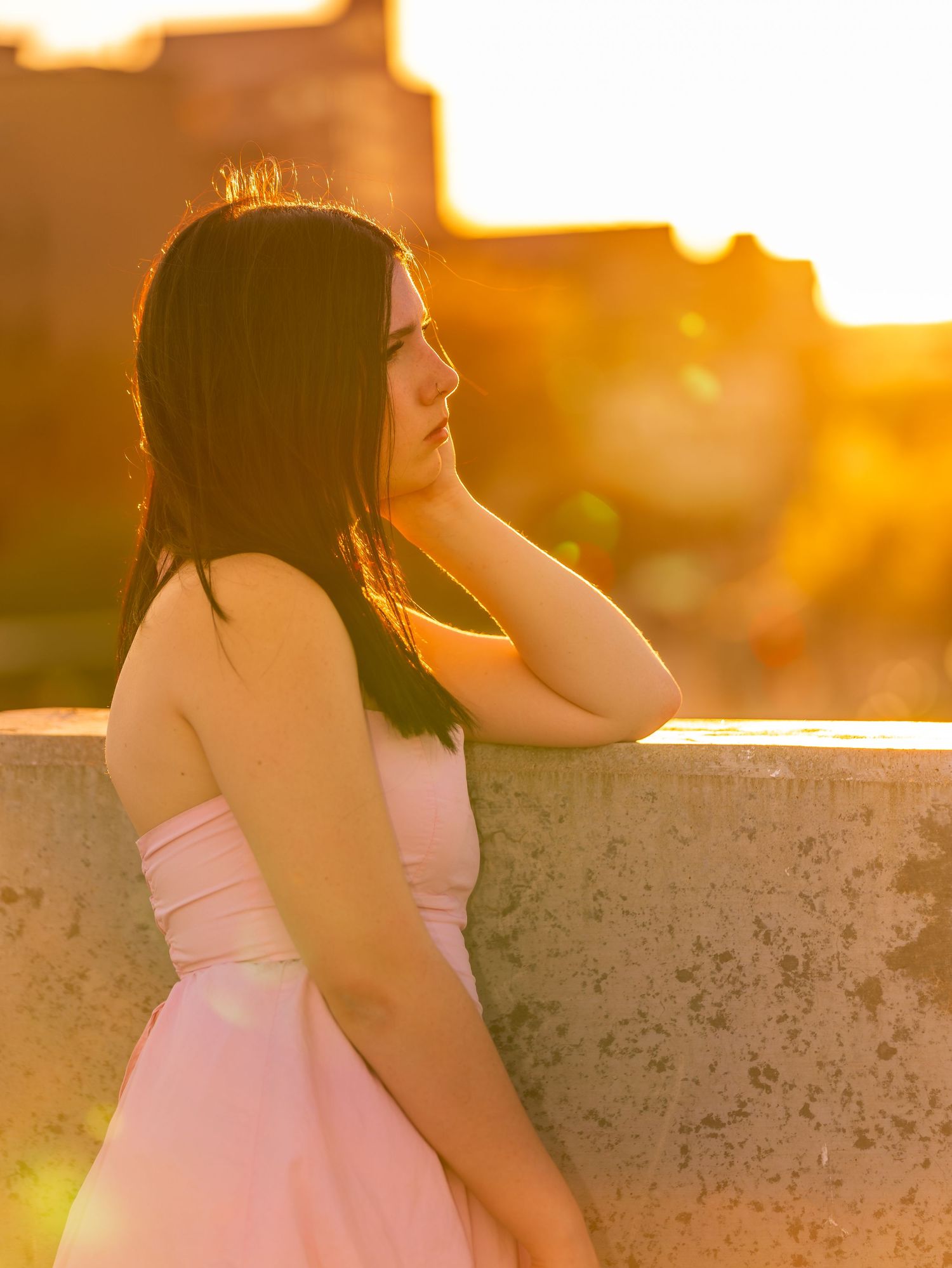 Young woman in pink dress leaning on stone wall bathed in warm sunset glow.