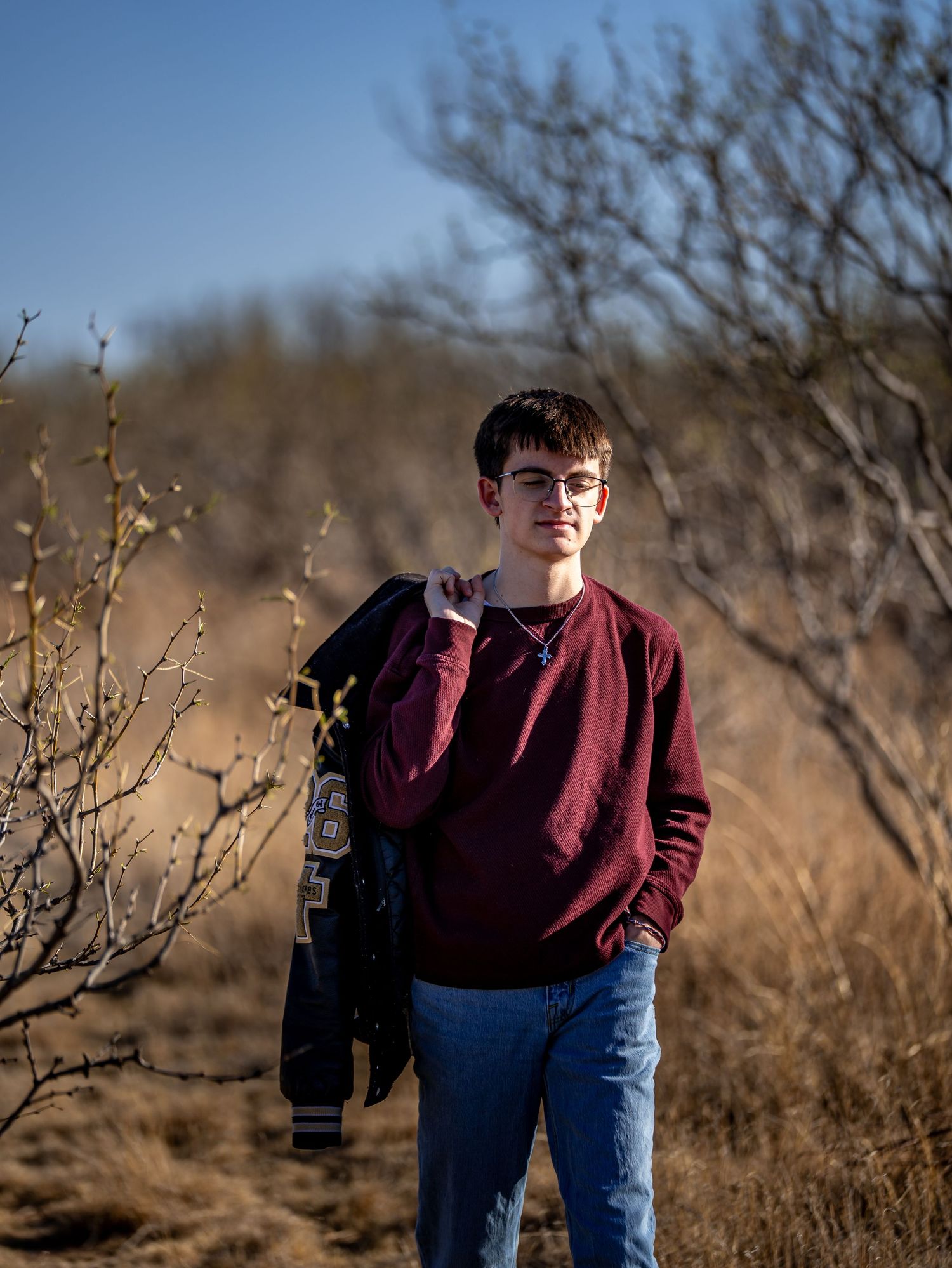 Young man in maroon sweater carrying a letterman jacket among dry brush in a winter landscape.