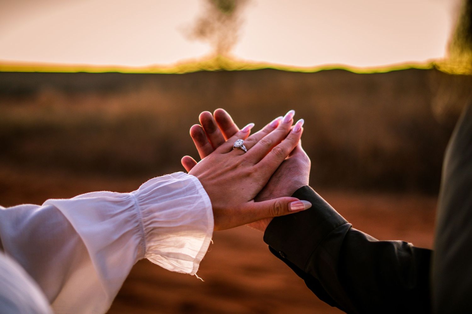 Close-up of couple holding hands displaying an engagement ring at golden hour outdoors.