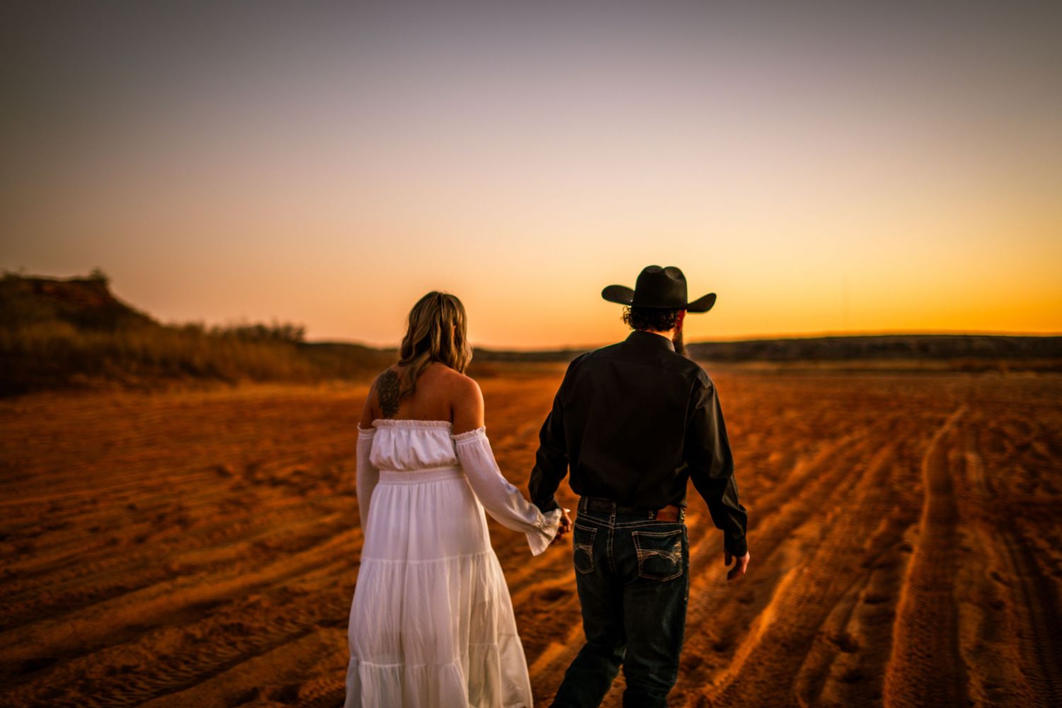 Couple holding hands walking into a golden sunset across a rustic red dirt field.
