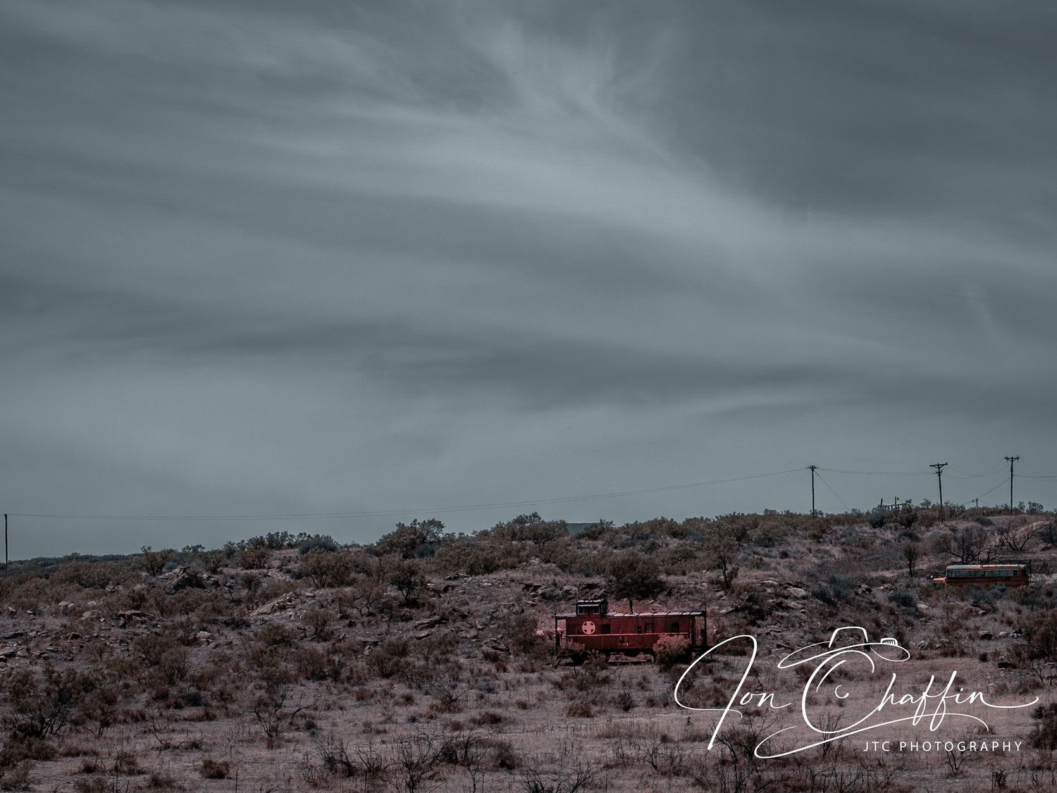 An abandoned red truck sits in a desolate desert landscape under a moody overcast sky with utility poles.