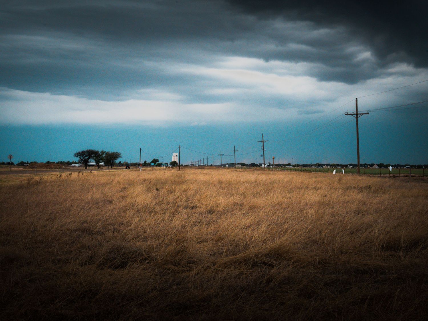 Dramatic stormy sky over a flat golden prairie with utility poles and distant structures.