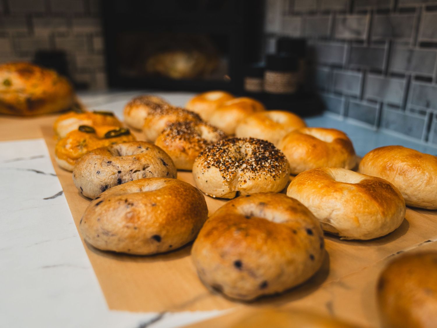 Freshly baked bagels with sesame and everything seasoning cooling on parchment paper in front of a stone fireplace.