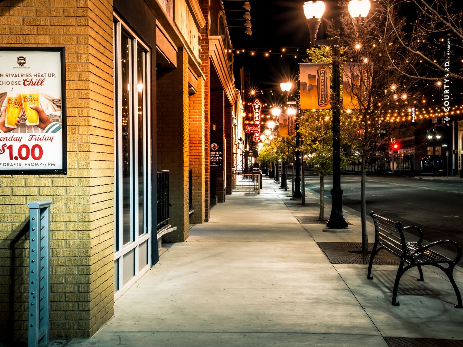 Nighttime urban sidewalk scene featuring string lights, park benches, and a Sonic chili advertisement on a brick building.