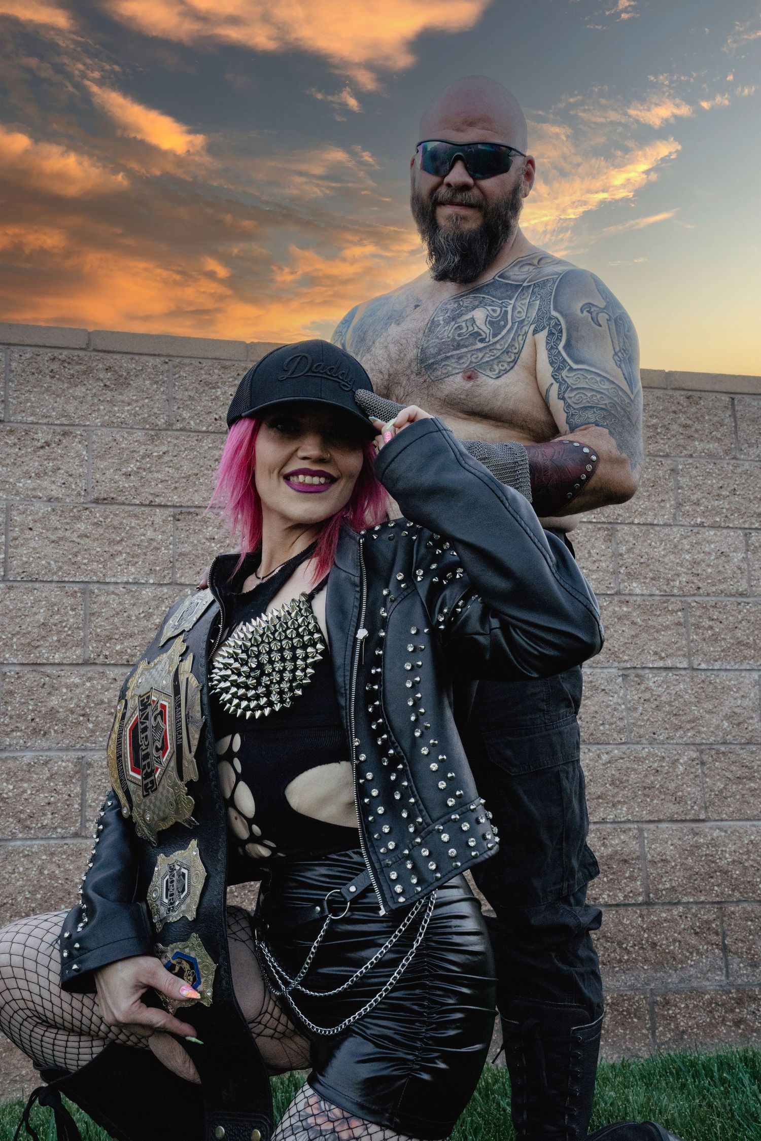 Punk rock couple in studded leather outfits posing at sunset, woman with pink hair smiling.