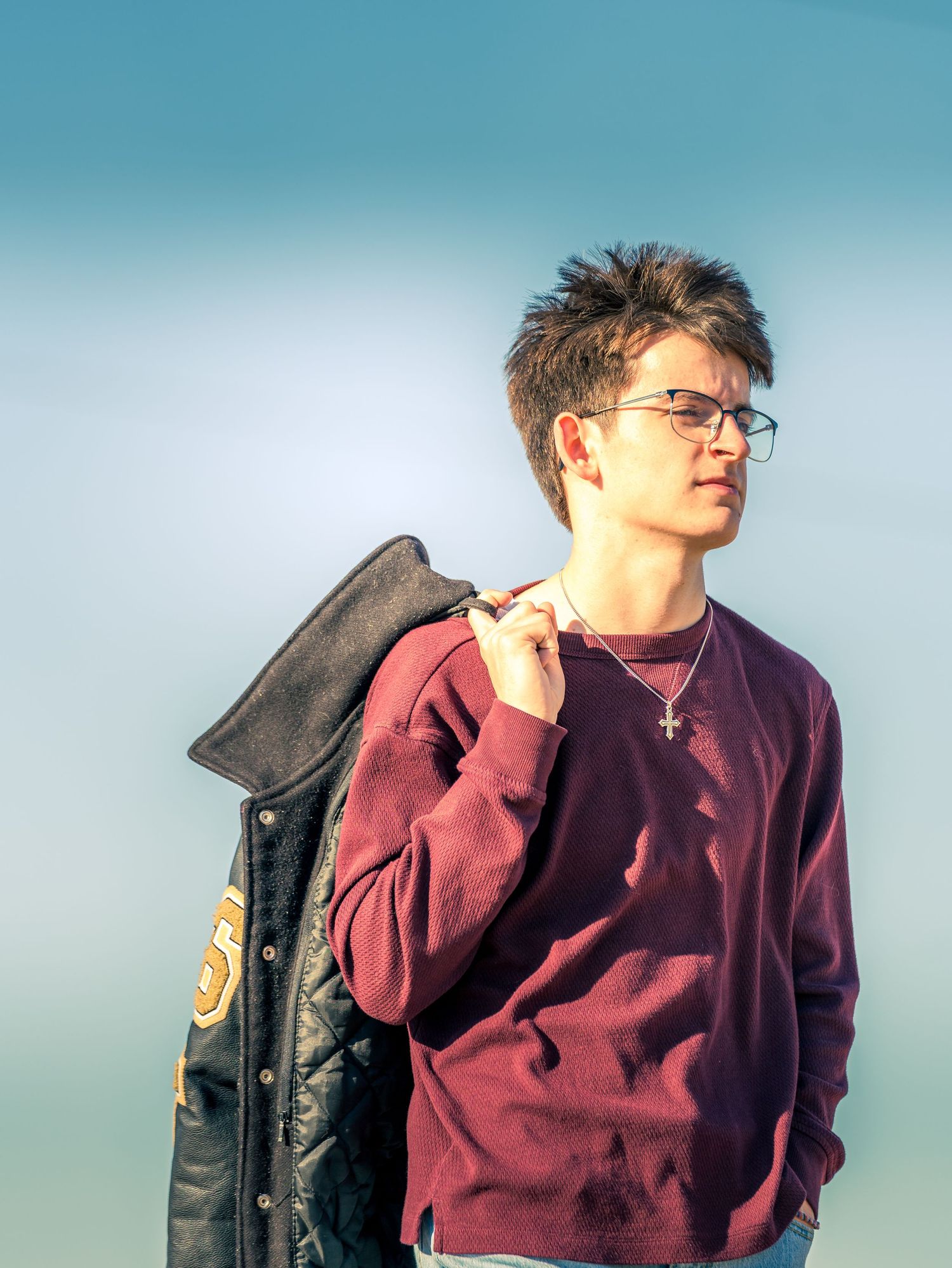 Young man in maroon sweater holding a letterman jacket over his shoulder against a blue sky.