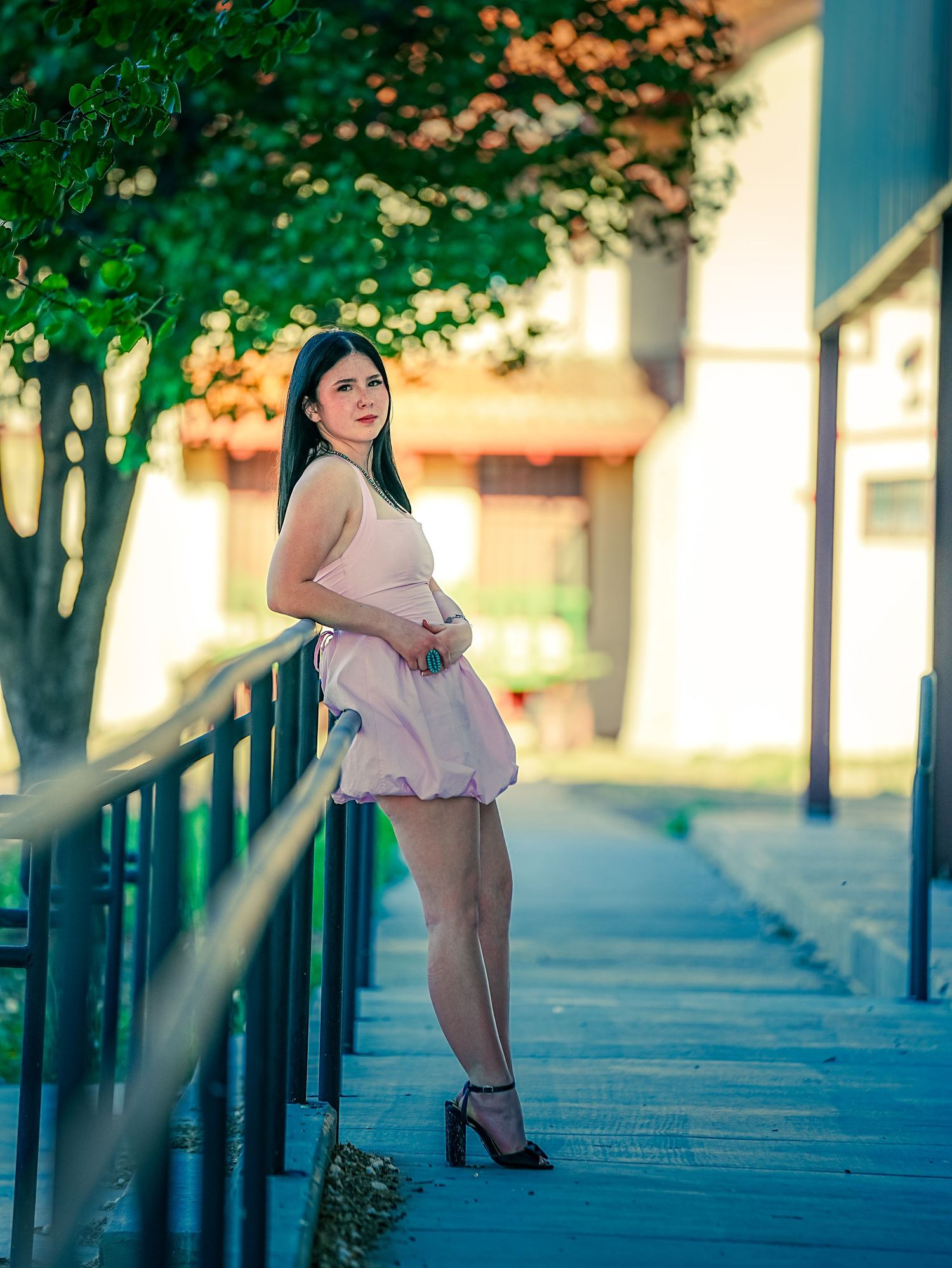 Young woman in pink bubble dress posing by railing with trees and building at golden hour.