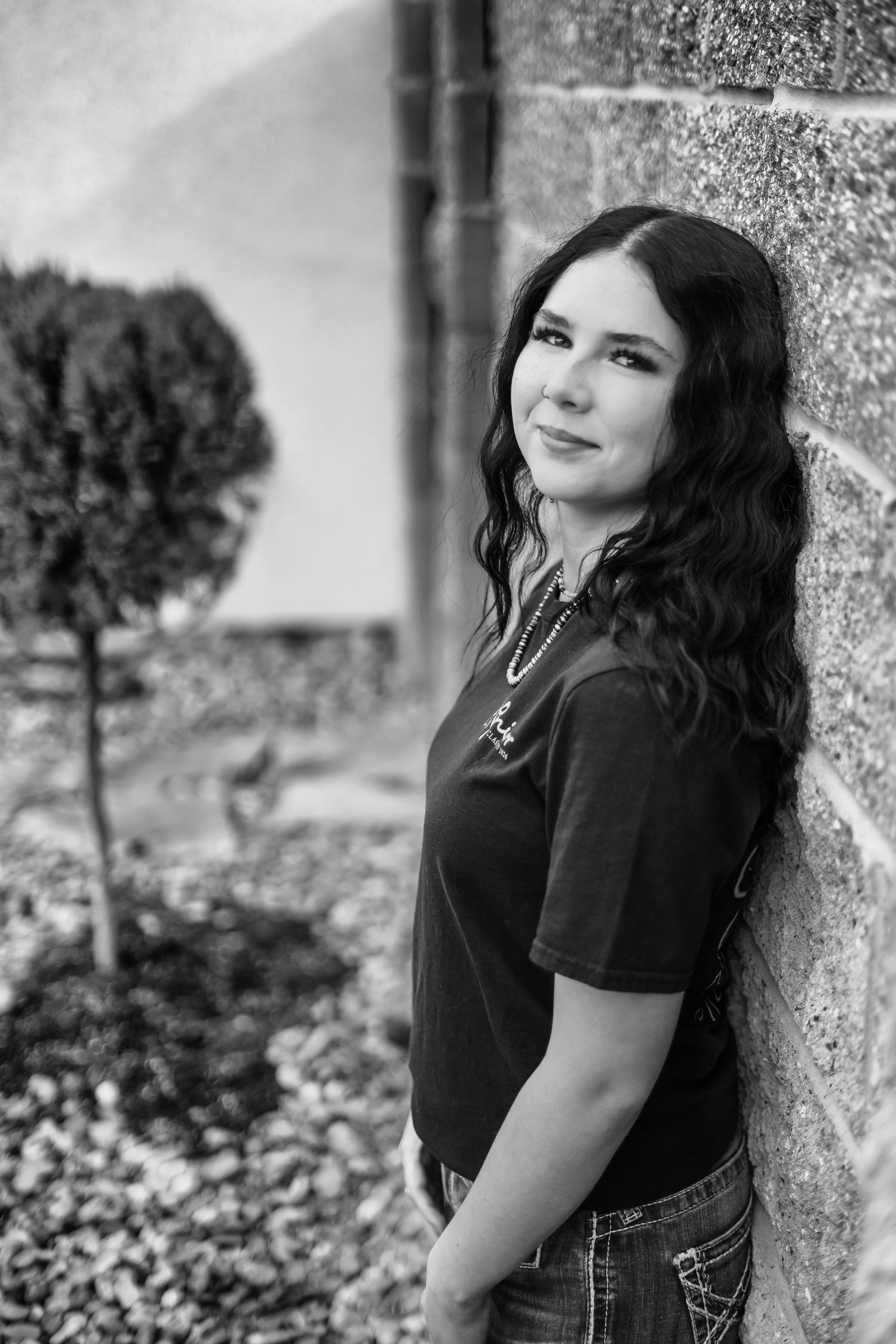 Black and white portrait of a young woman with wavy dark hair smiling while leaning against a stone wall.