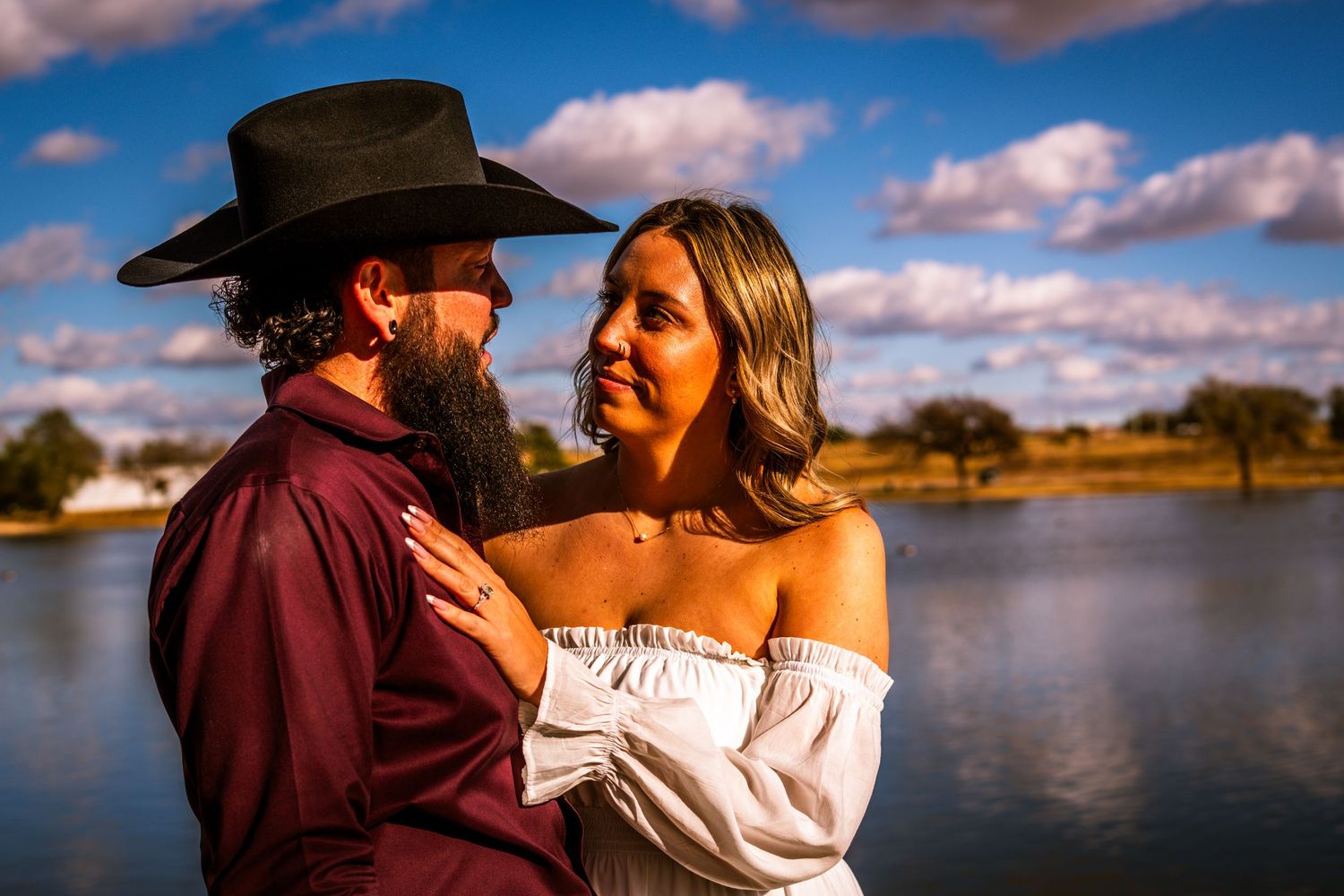 Couple embracing near a lake, woman in white off-shoulder dress and man in black cowboy hat under blue sky.