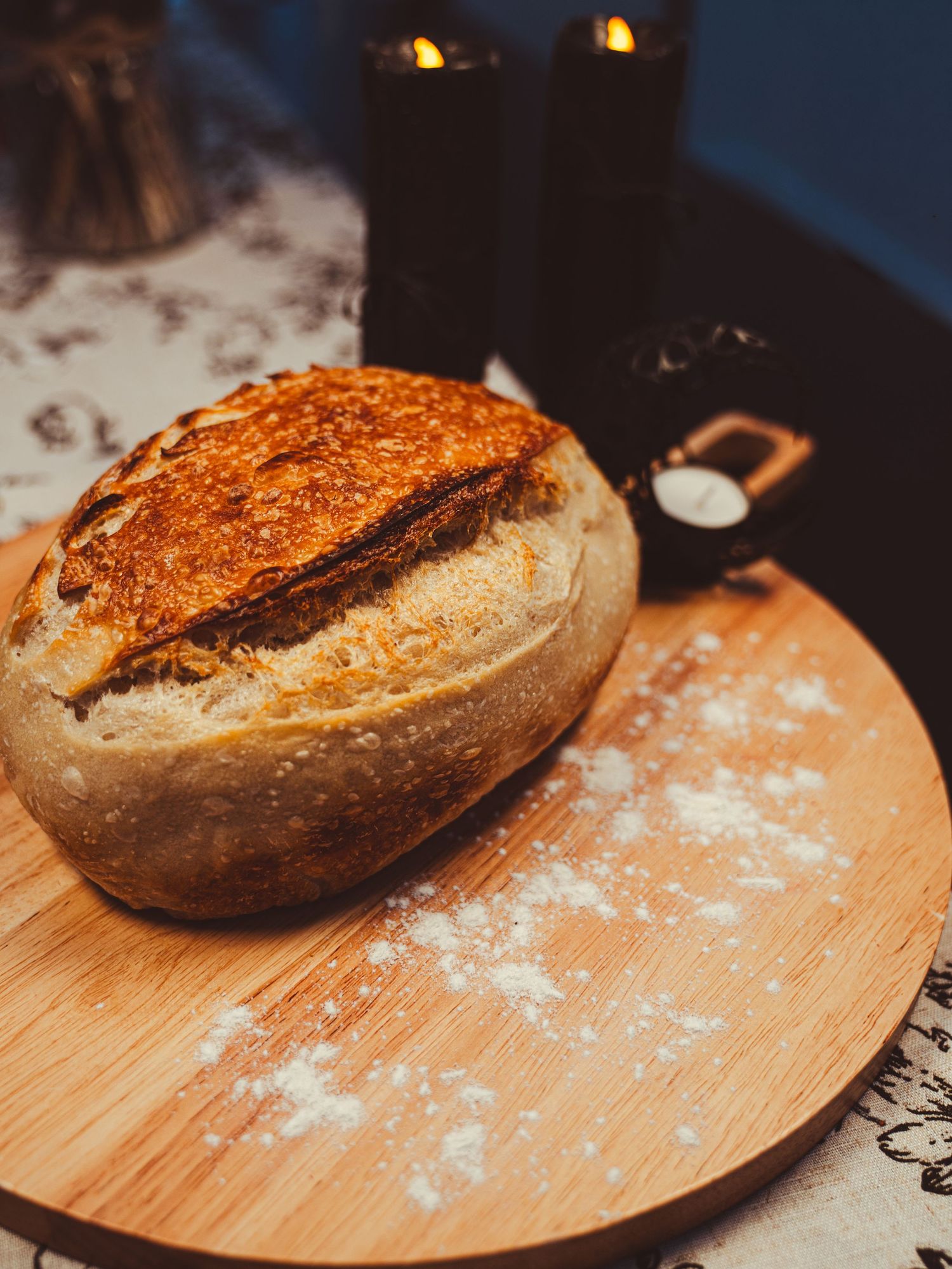 Rustic sourdough bread loaf on wooden board dusted with flour, surrounded by candles in moody lighting.