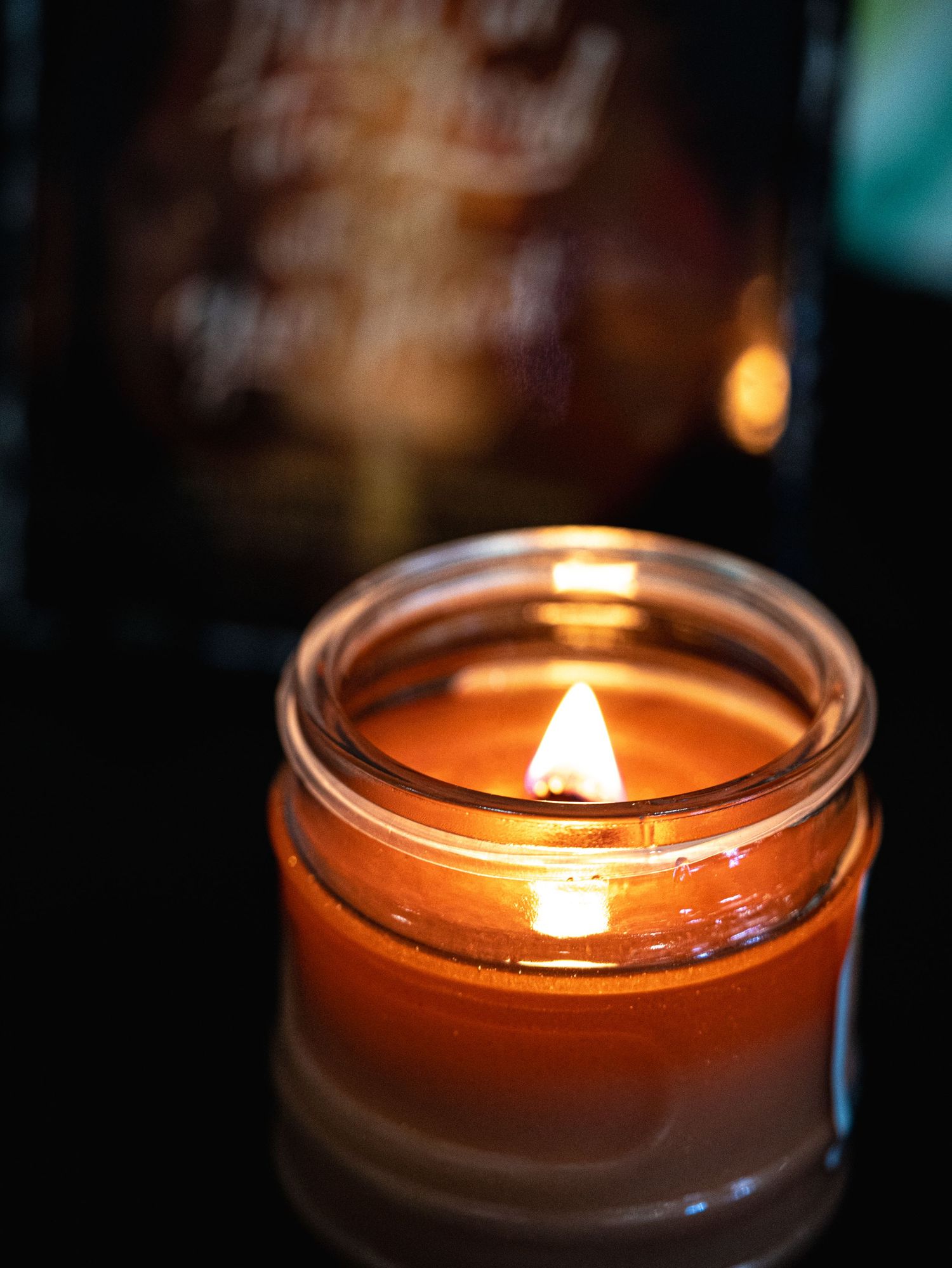 A glowing amber jar candle with a warm flame burns against a dark background with a blurred bottle behind it.