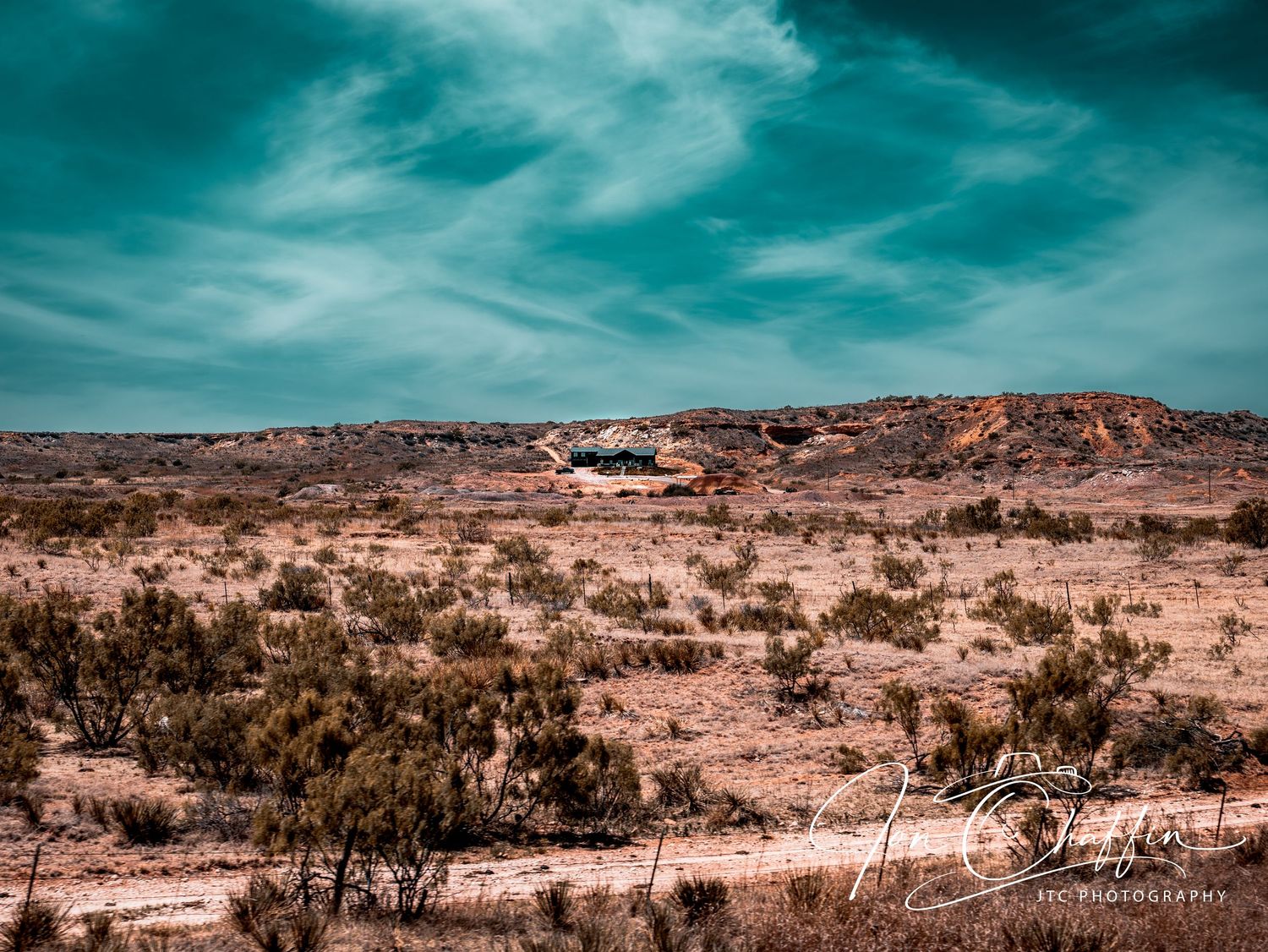 Desert scrubland with a lone building under a vivid teal sky in arid terrain.