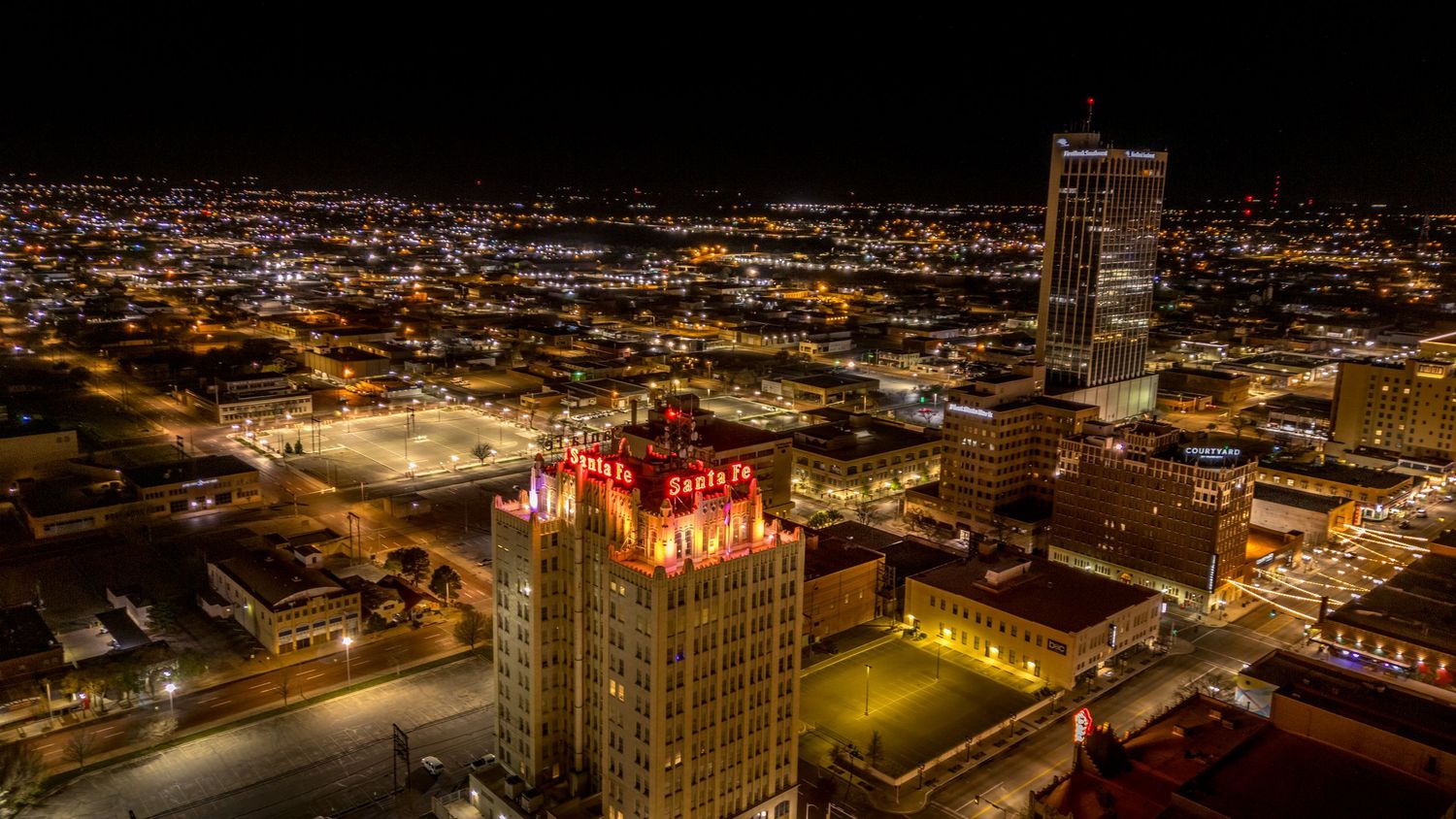 Aerial night view of downtown with the Santa Fe building glowing in warm gold light amid city streets and skyscrapers.