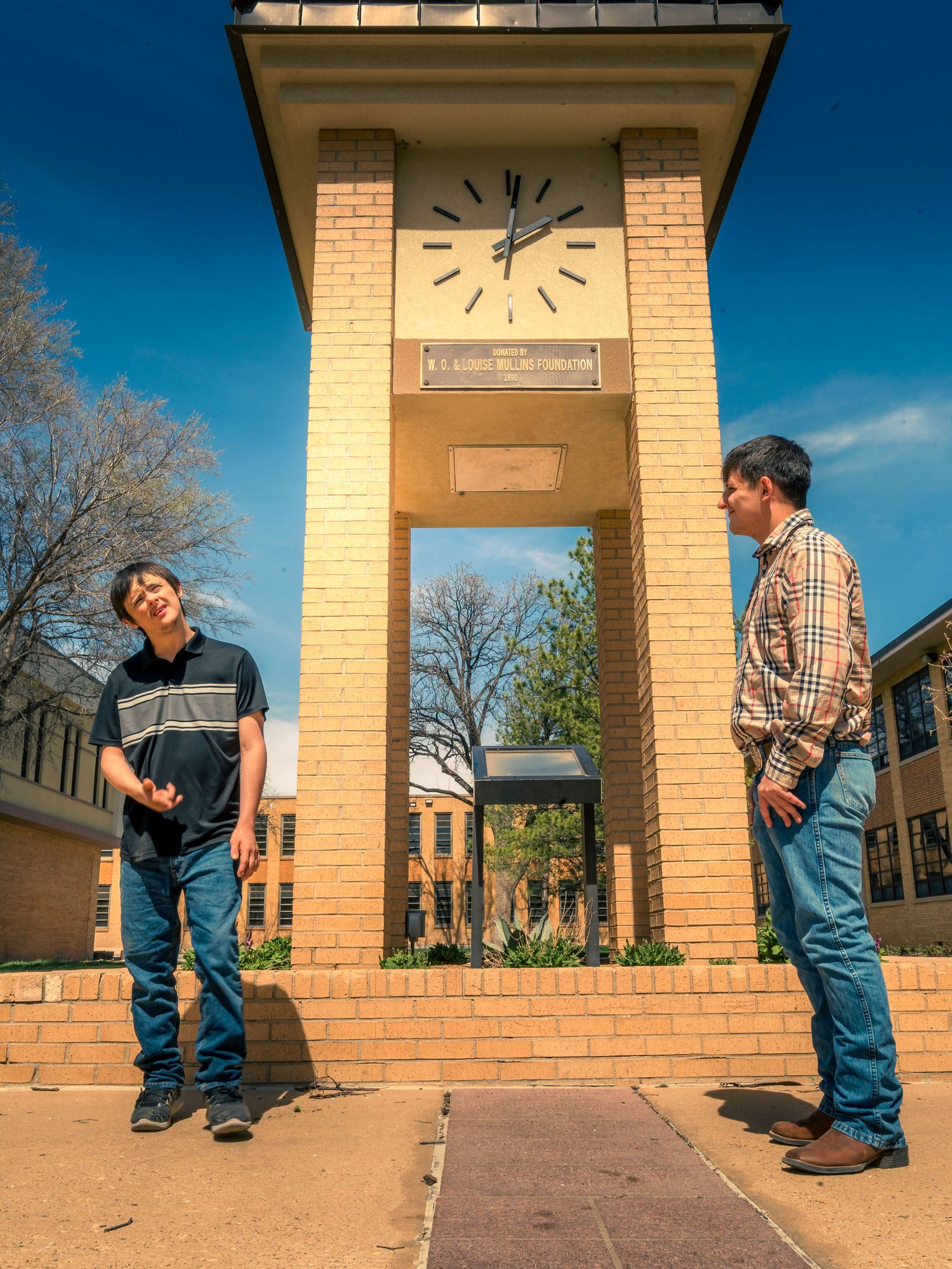 Two young men standing apart by a brick clock tower on a sunny college campus.