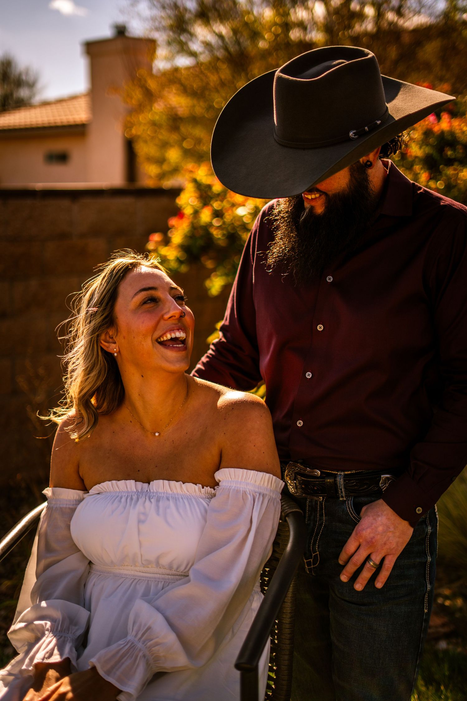 Laughing woman in white off-shoulder dress shares a joyful moment with bearded man in black cowboy hat at sunset.