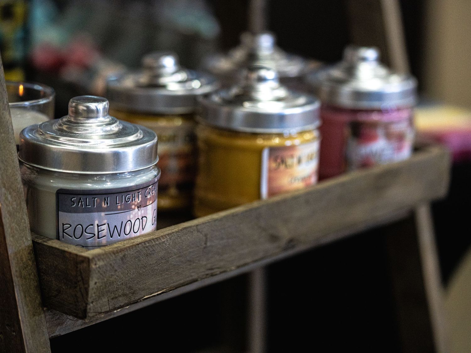 Rustic wooden shelf displaying decorative scented candle tins, including a Rosewood fragrance candle with silver lids.