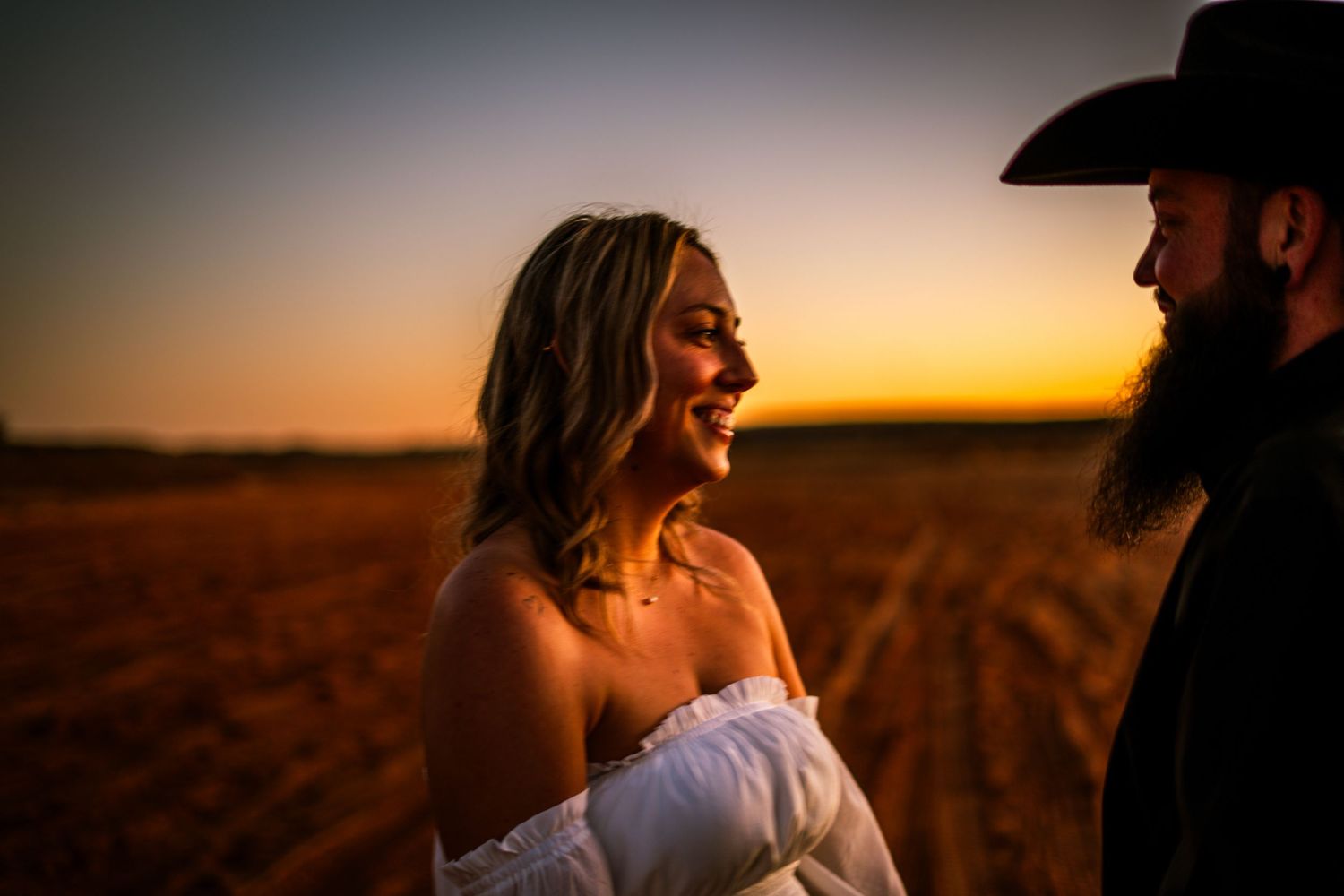 Smiling bride in white dress with bearded groom in black cowboy hat at sunset.