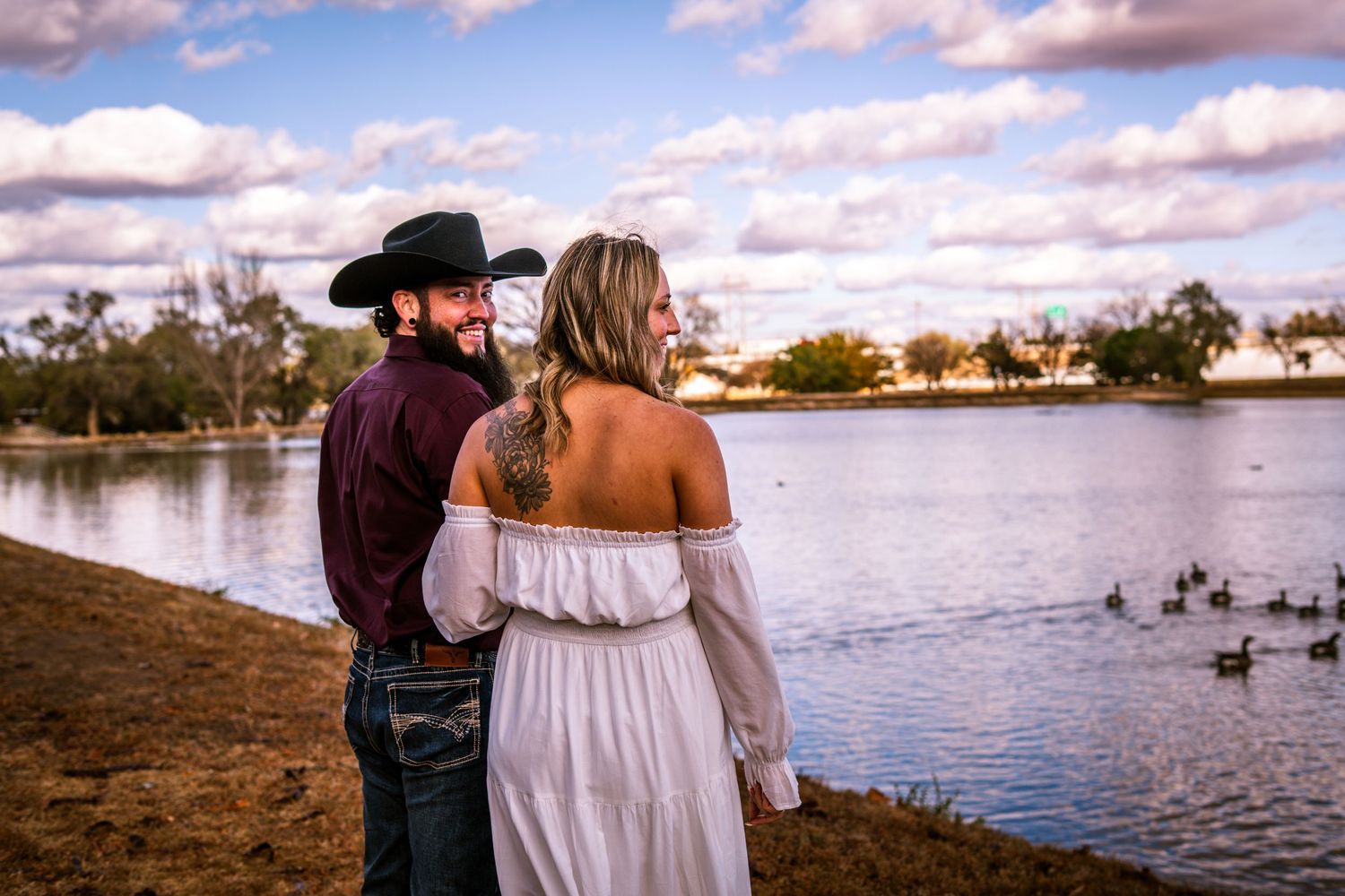 Couple in white dress and cowboy hat standing by a lake watching ducks under a dramatic cloudy sky.